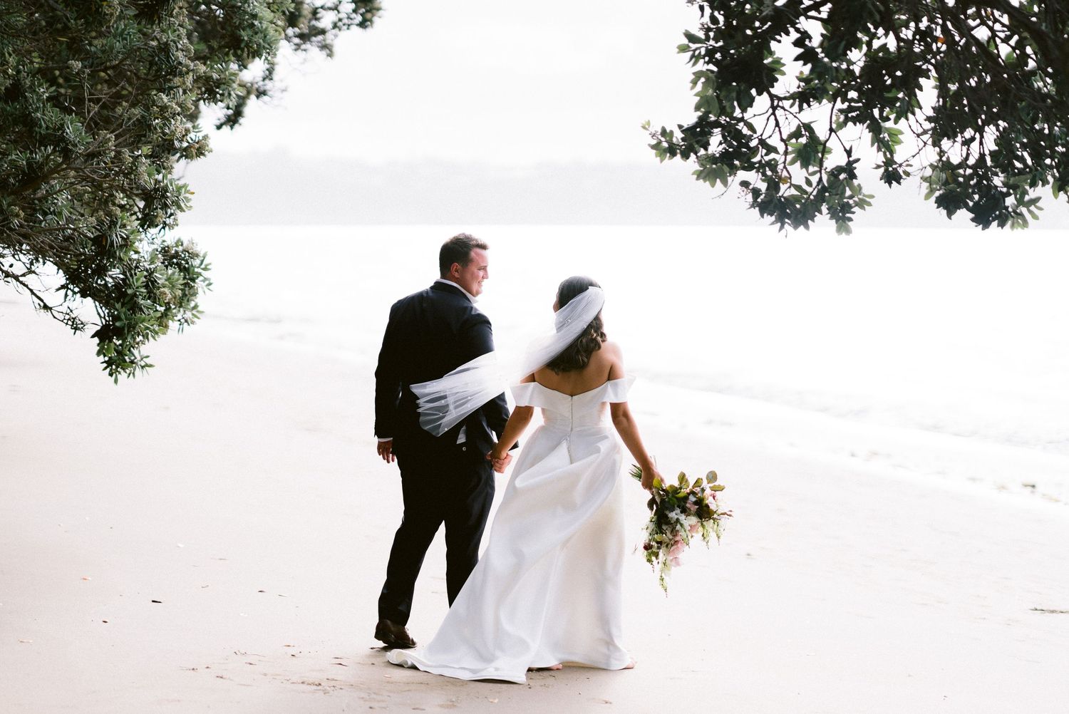 Couple walks together on beach holding hands with floral bouquet during wedding celebration.