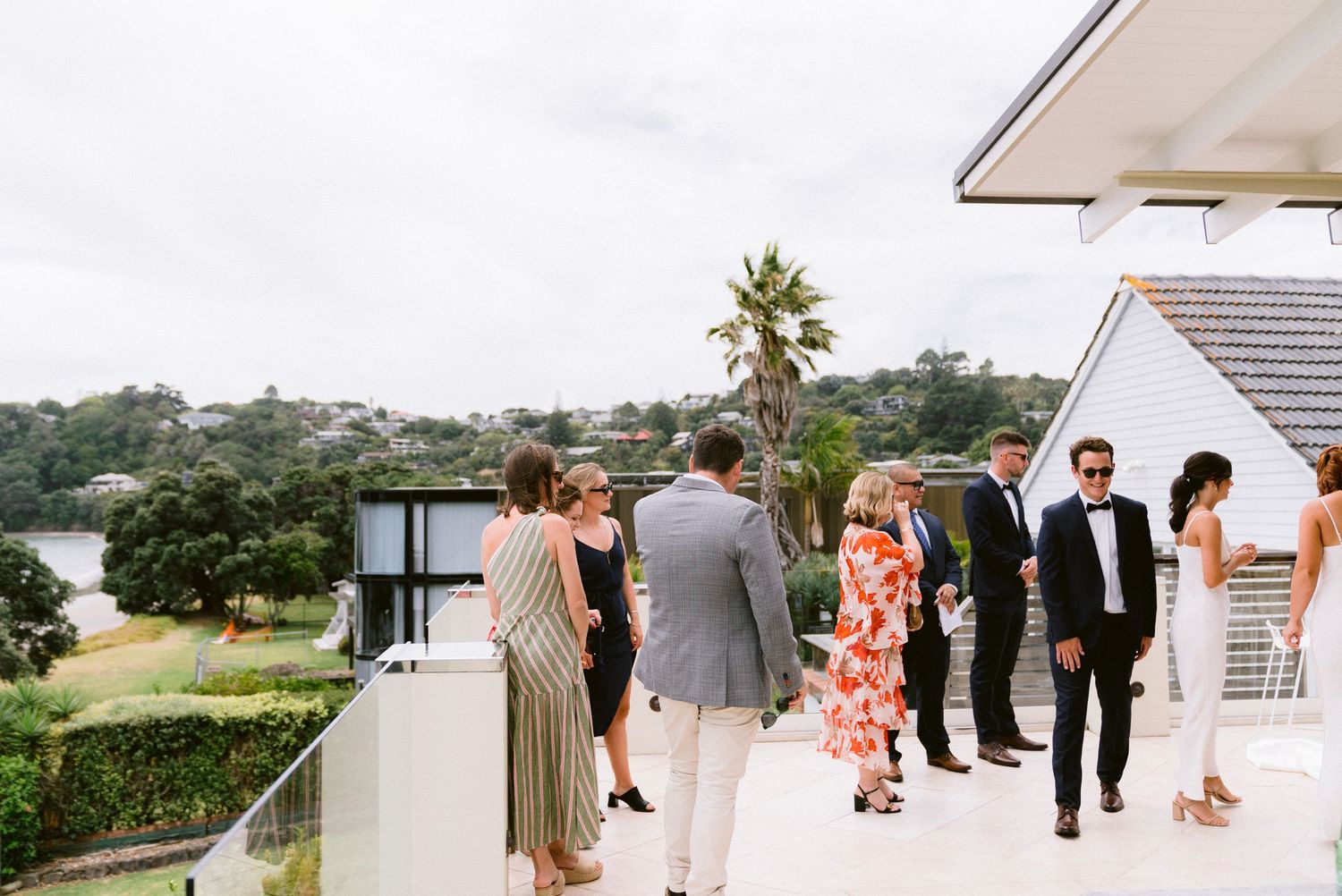 Wedding guests mingle on an outdoor terrace overlooking a scenic waterfront view.