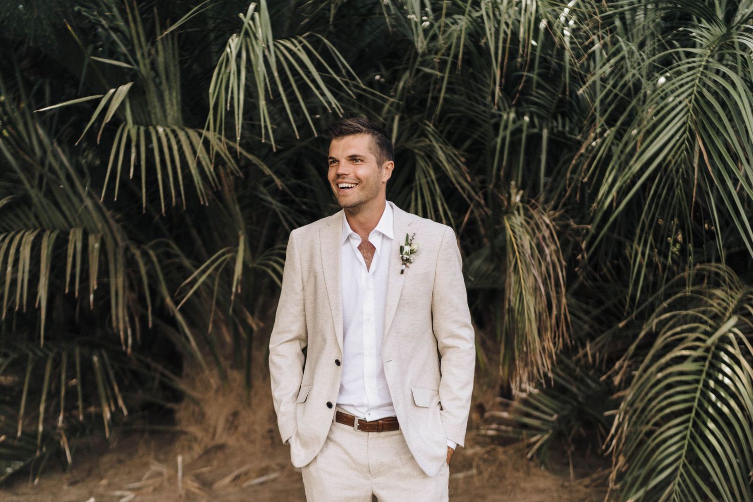 Wedding party members pose against tropical palm background in light colored attire.