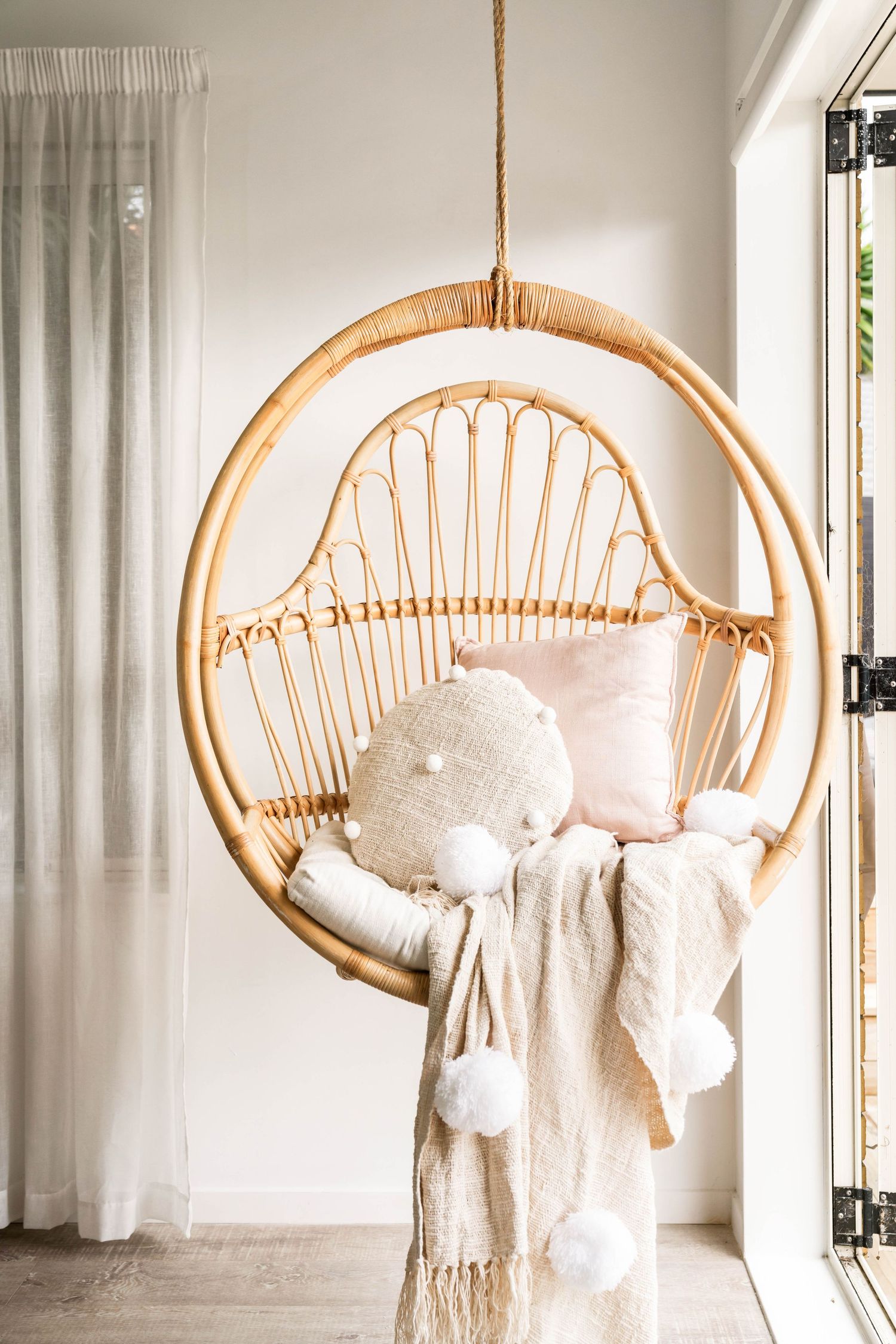 Hanging rattan egg chair decorated with cozy pillows and throw blankets in a bright corner space.