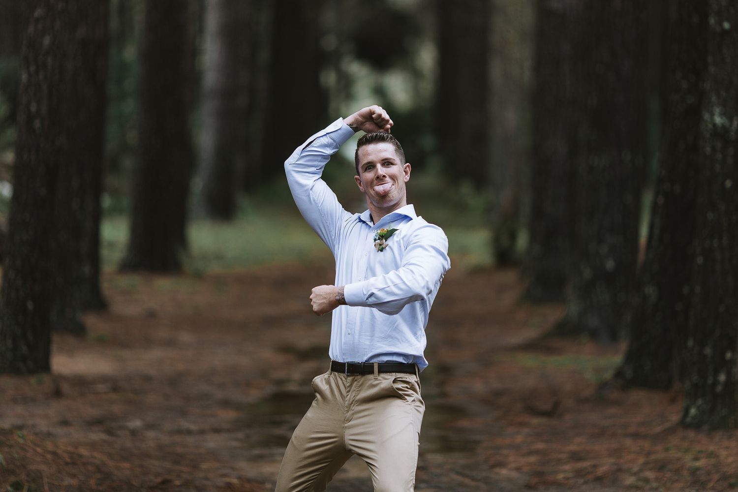 Well-dressed person strikes a joyful pose while walking along a scenic forest path lined with tall pine trees in soft light.