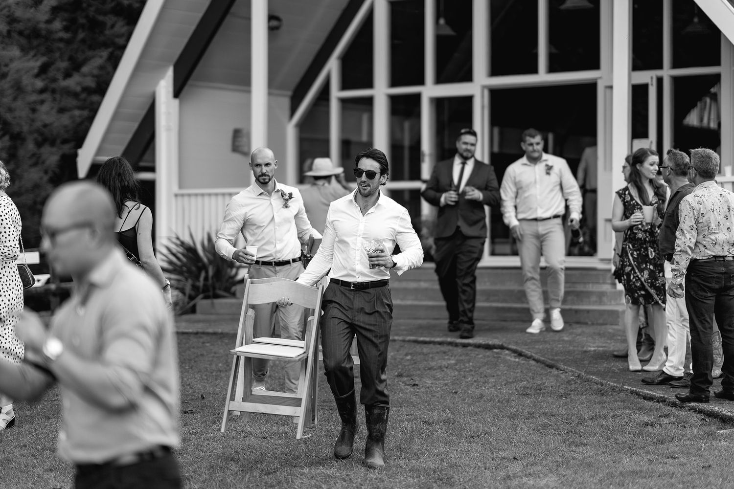 Black and white image showing wedding guests mingling and waiting outdoors near white Adirondack chairs at an event.