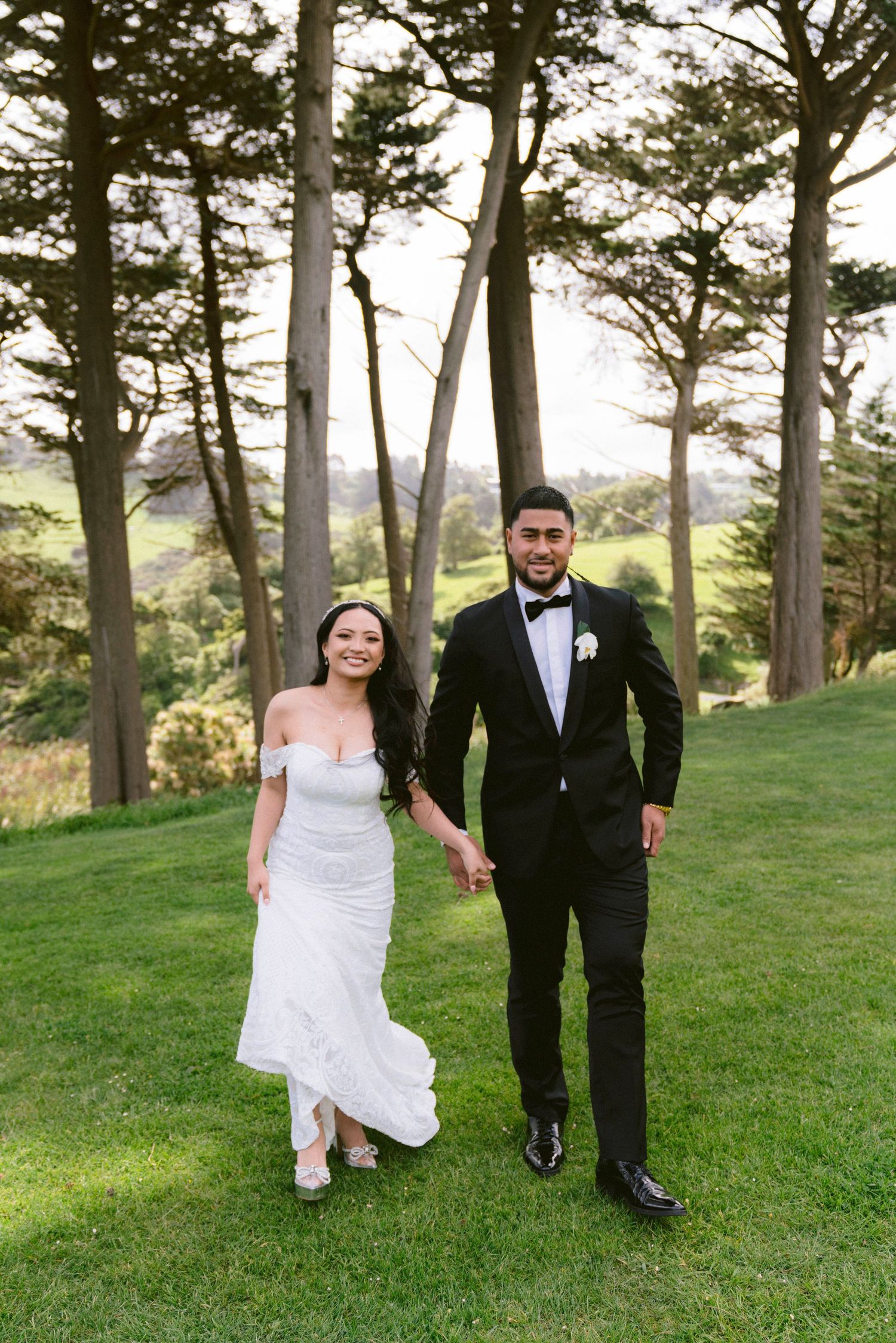 Newlywed couple strolls through scenic pine grove on grassy slope in formal wedding attire.