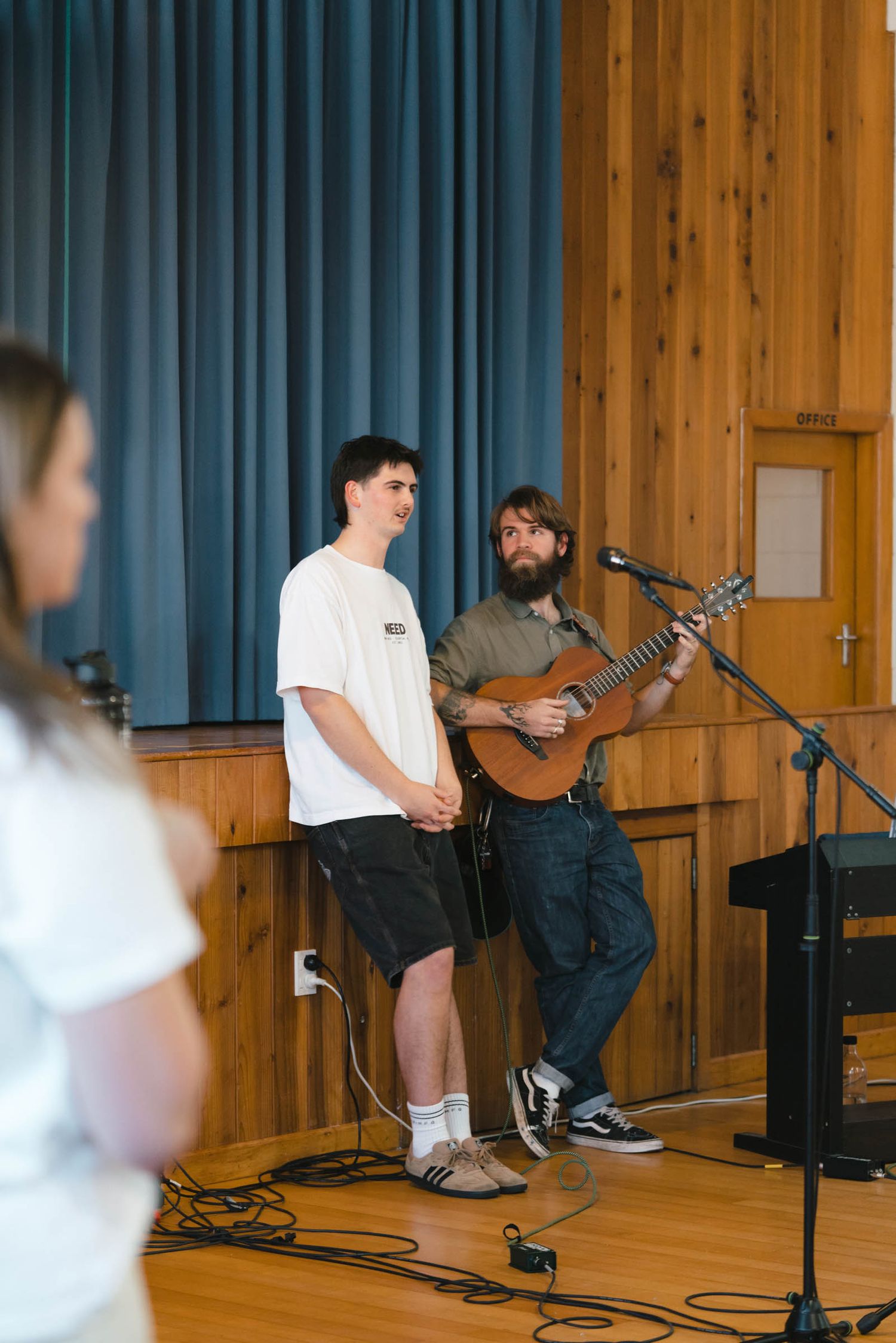 Musicians performing on stage with acoustic guitar in a wood-paneled room with blue curtains.