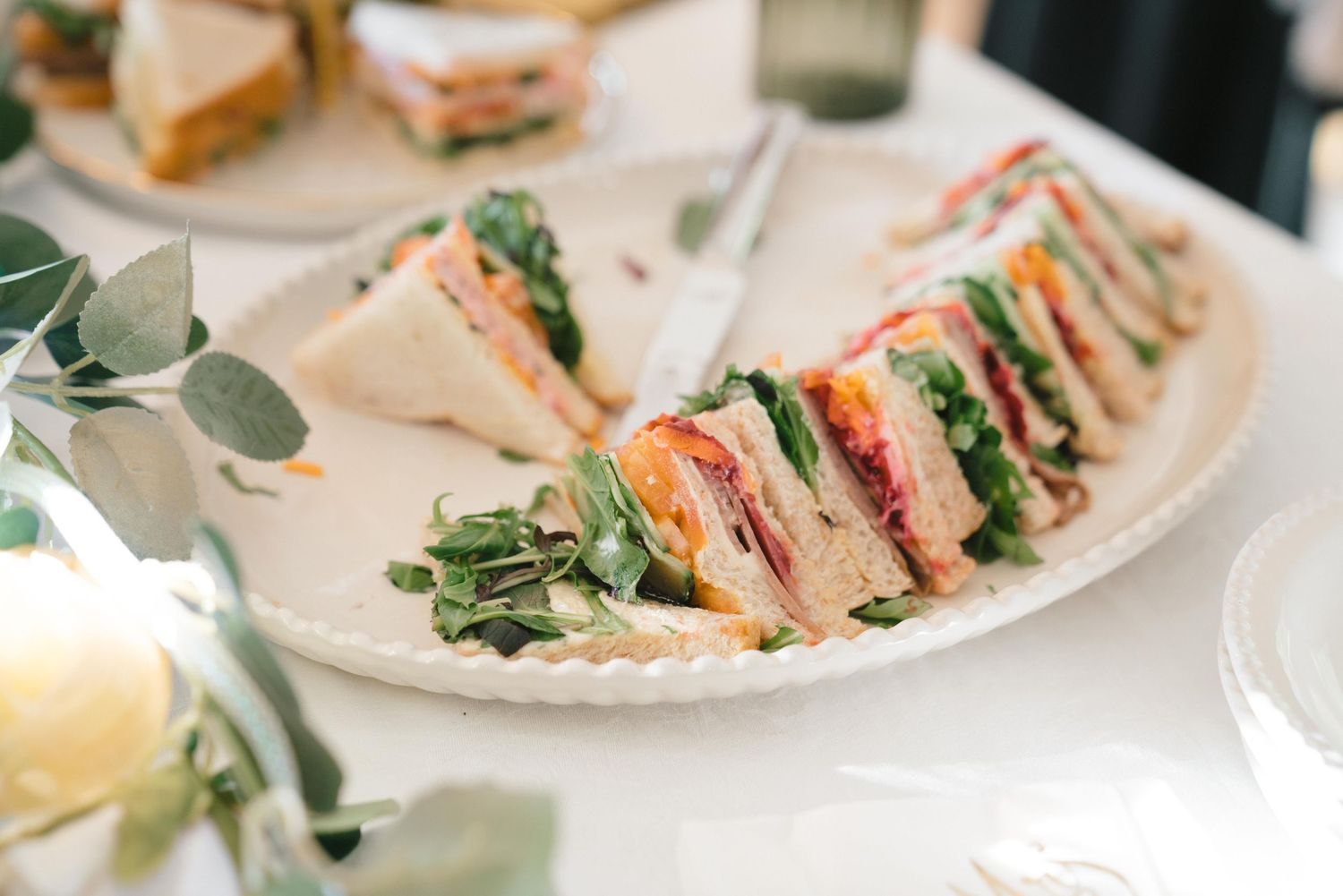Fresh club sandwiches arranged on a white plate with eucalyptus garnish for catered event.