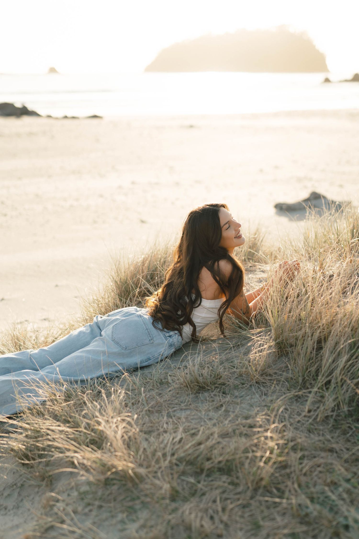 Figure reclines peacefully in light clothing among beach grass at sunset.