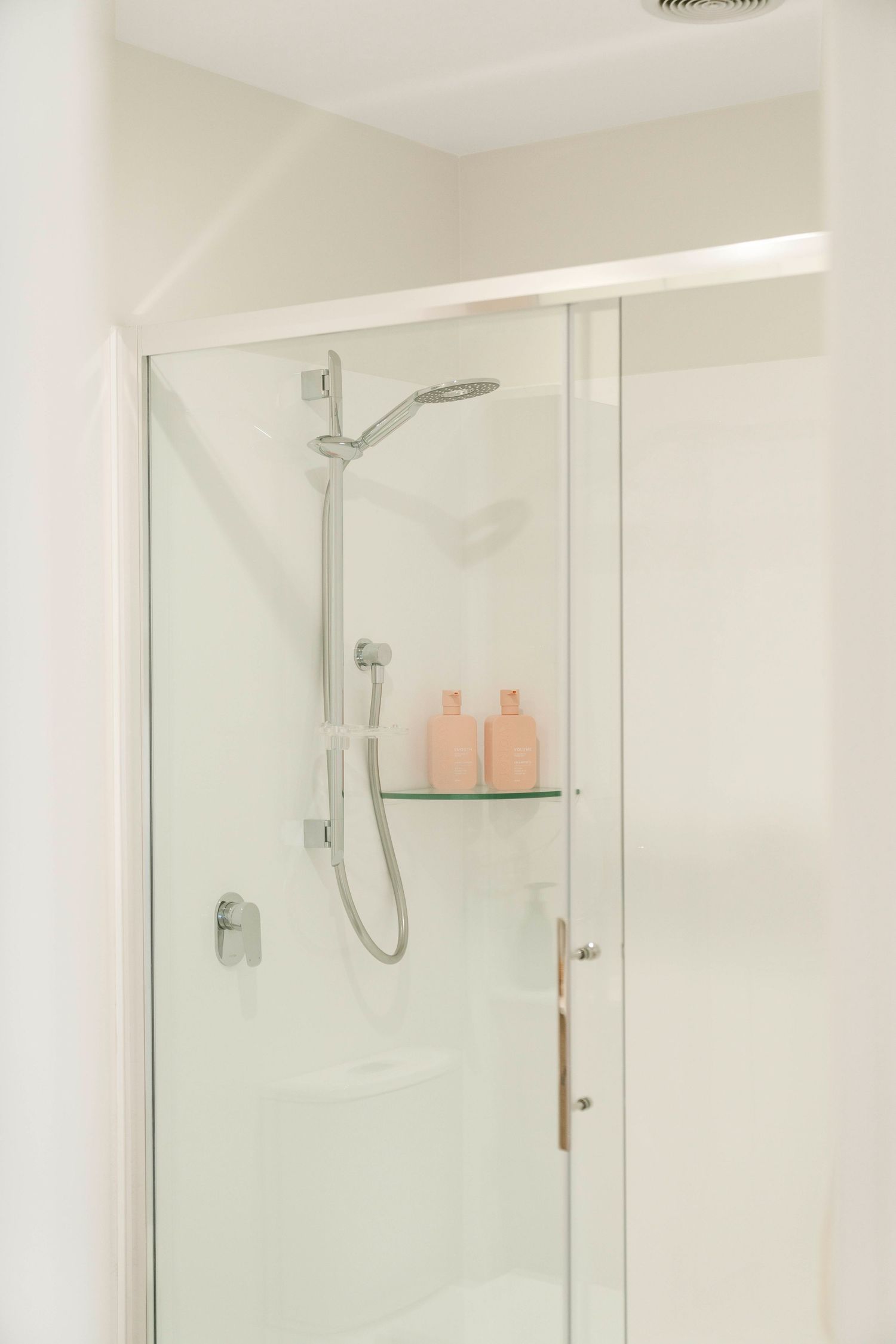 Clean white shower enclosure with glass door and pink toiletry bottles on shelf.