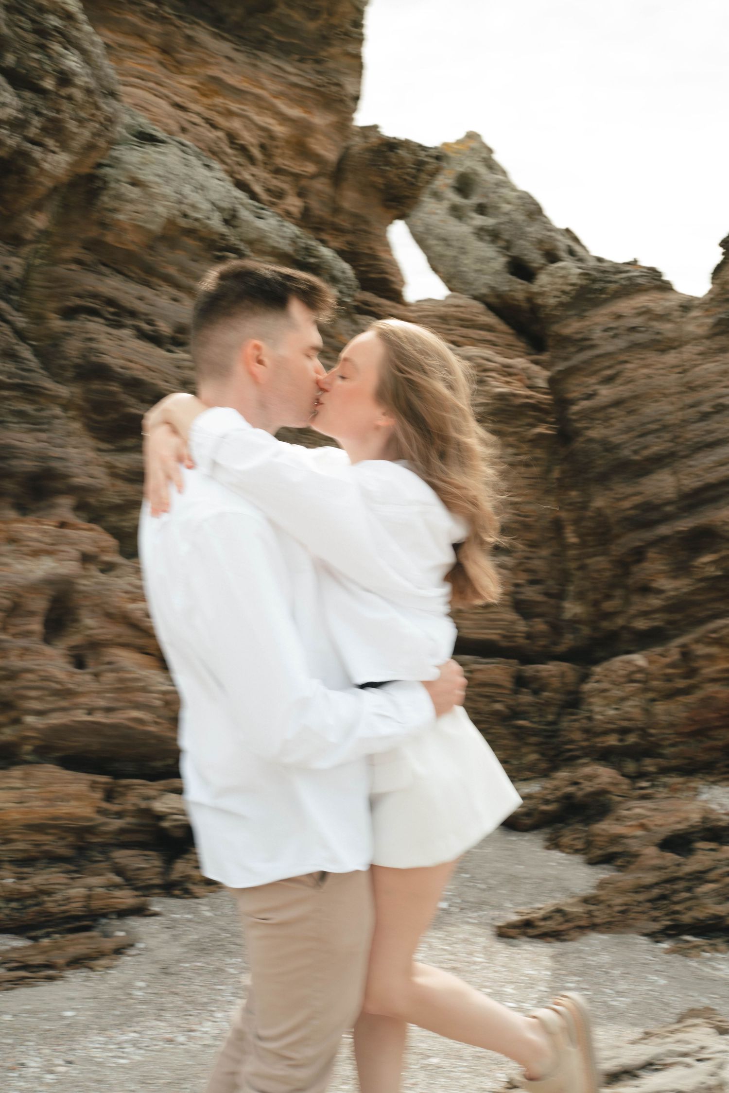 Two people in white embrace and dance romantically against a backdrop of dramatic coastal rocks.