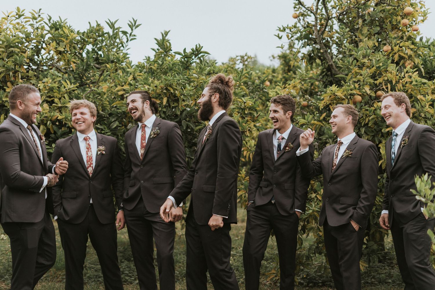 Groomsmen in dark suits share candid moments and laughter together during outdoor wedding photo session at garden venue.