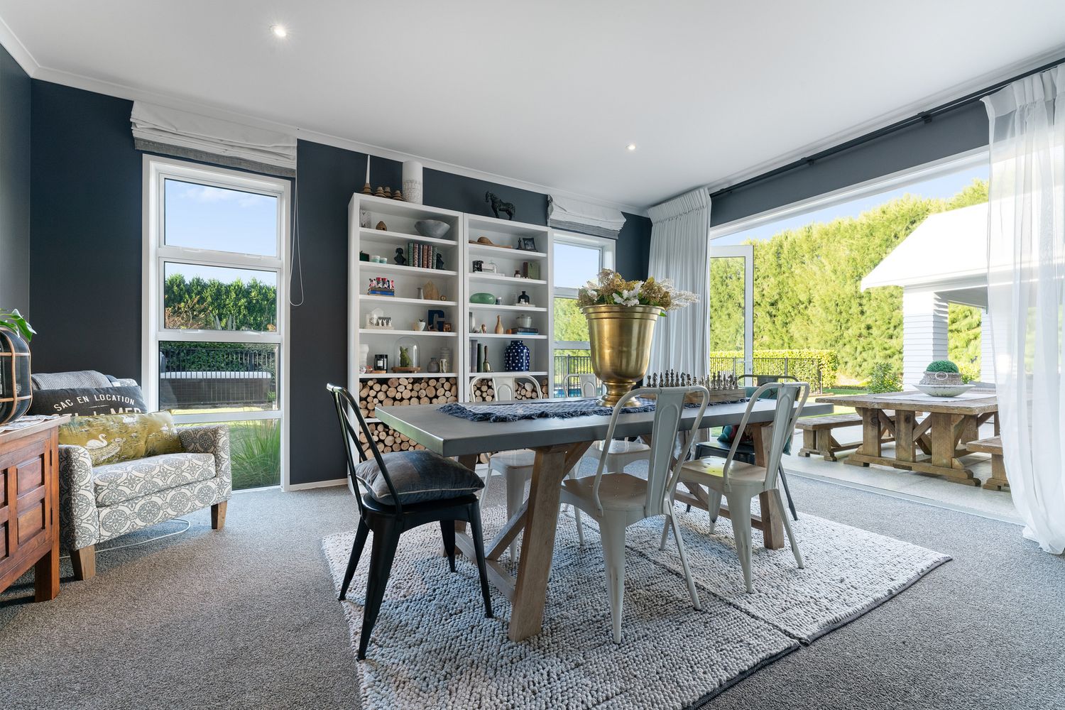 Contemporary dining room with navy walls, mixed seating, built-in shelving, and large windows overlooking the garden.