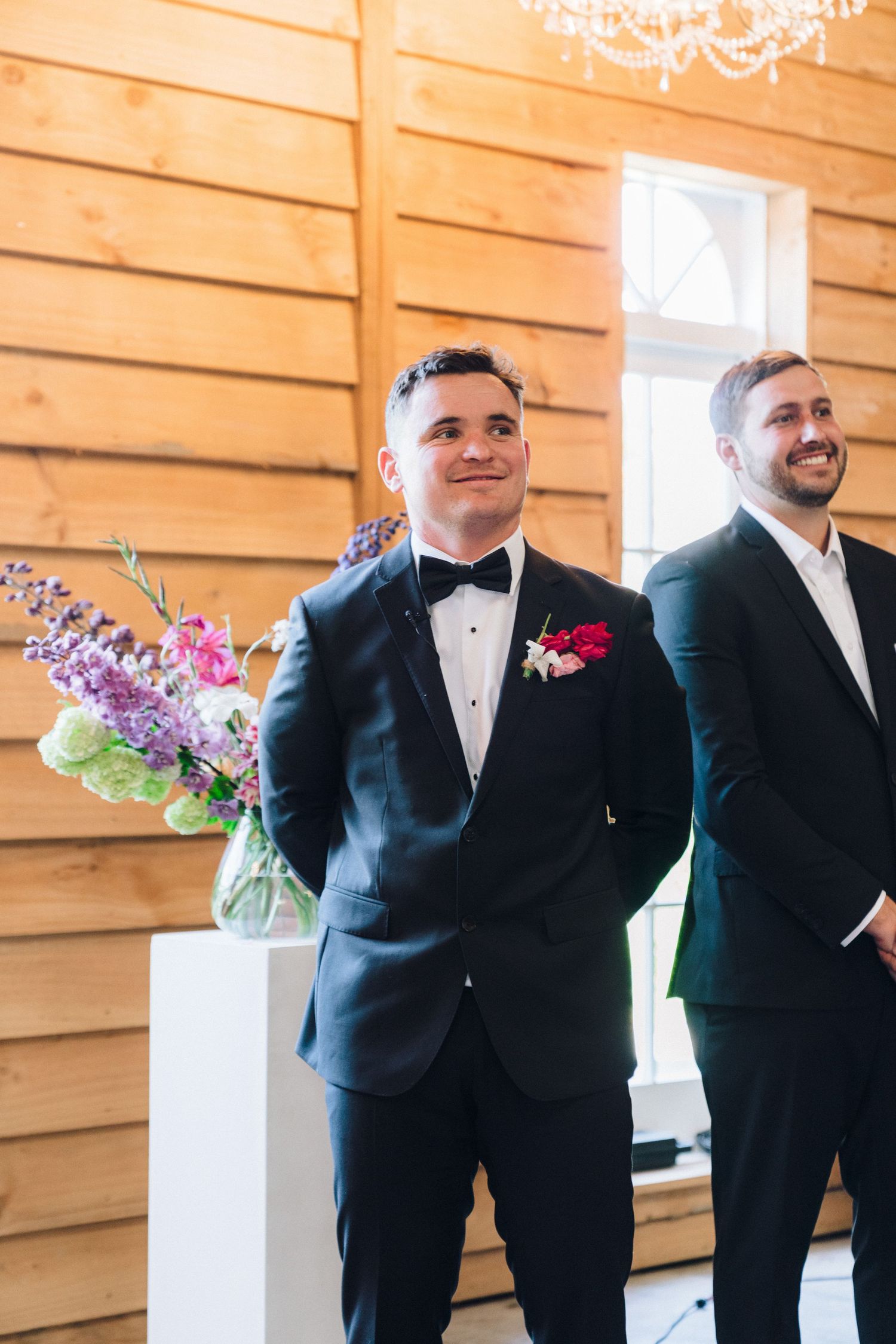 Two wedding attendants in black tuxedos stand together smiling during ceremony in rustic wood-paneled venue.