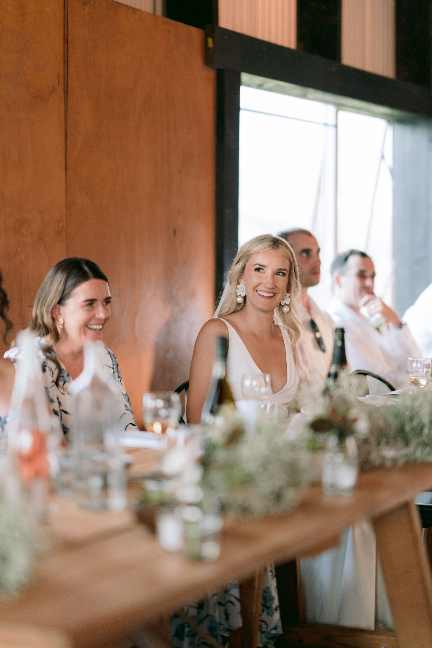 A candid scene at a wedding reception shows joyful guests laughing around a wooden dining table with floral decorations.