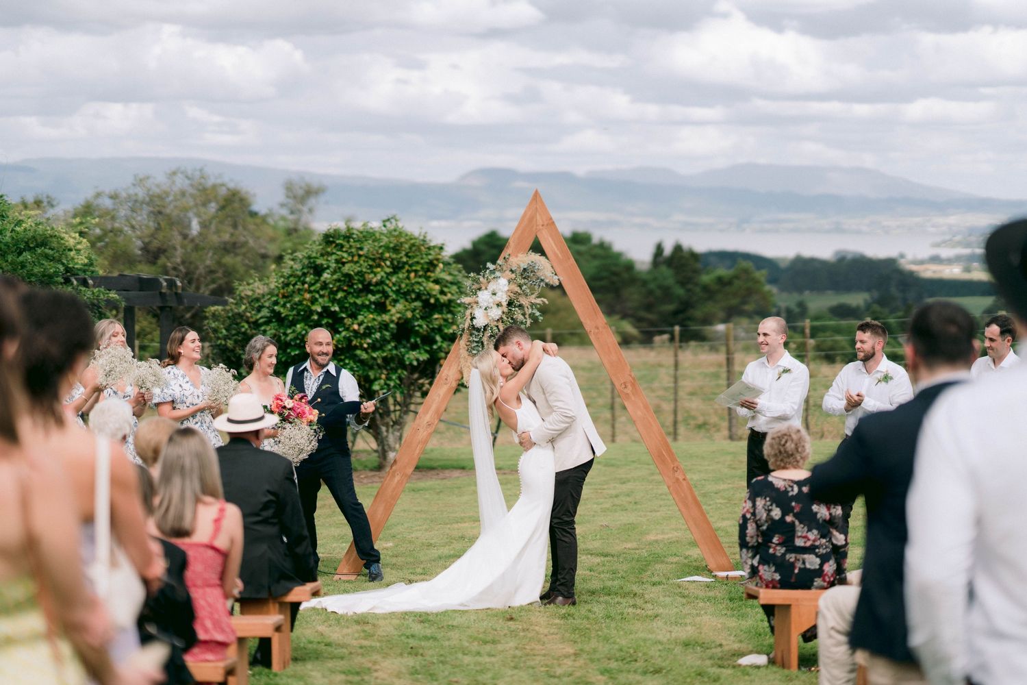 Wedding ceremony under triangular wooden arch with guests witnessing special moment.