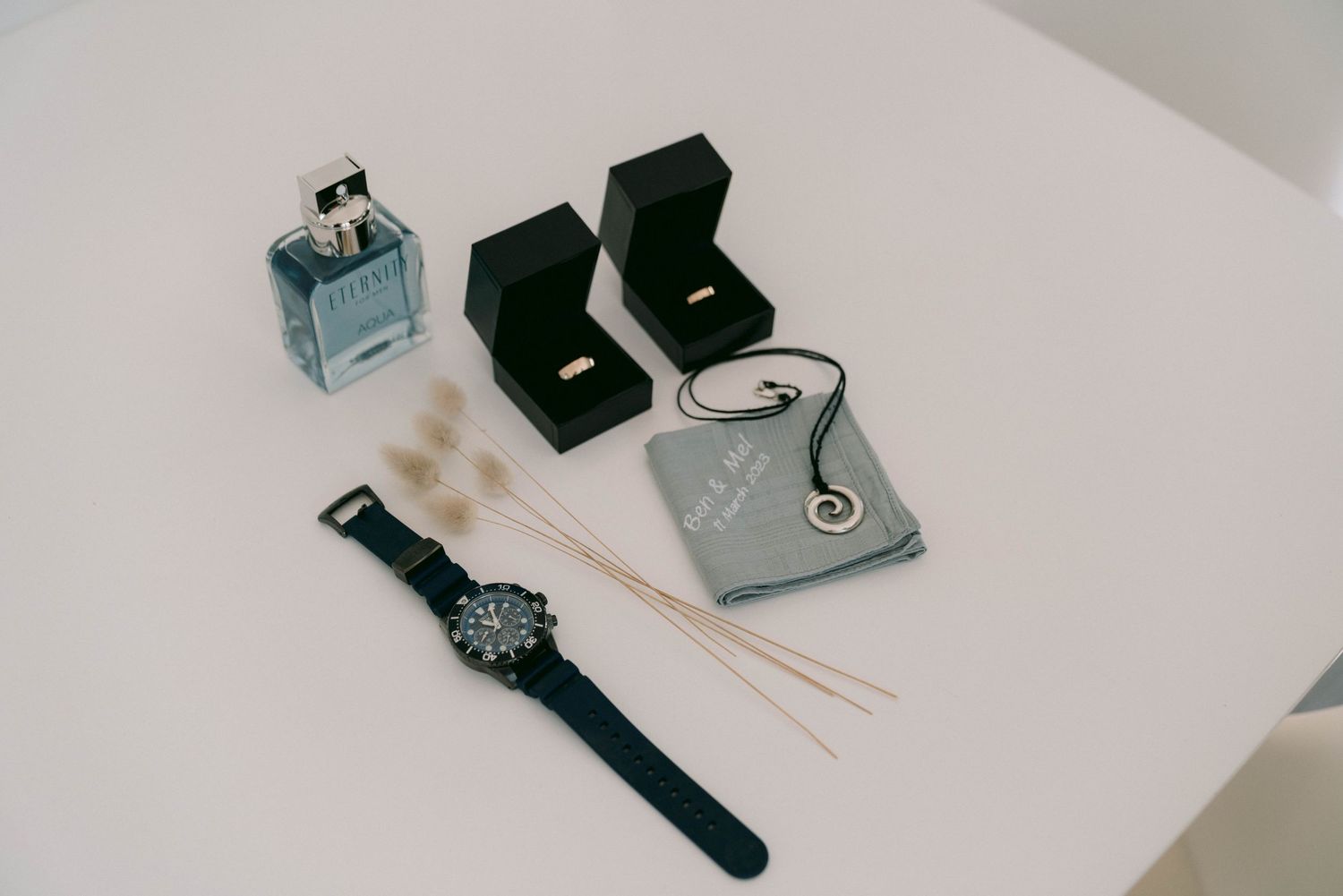 A flat lay arrangement of luxury accessories including a watch, perfume bottle, jewelry boxes and chopsticks on white surface.