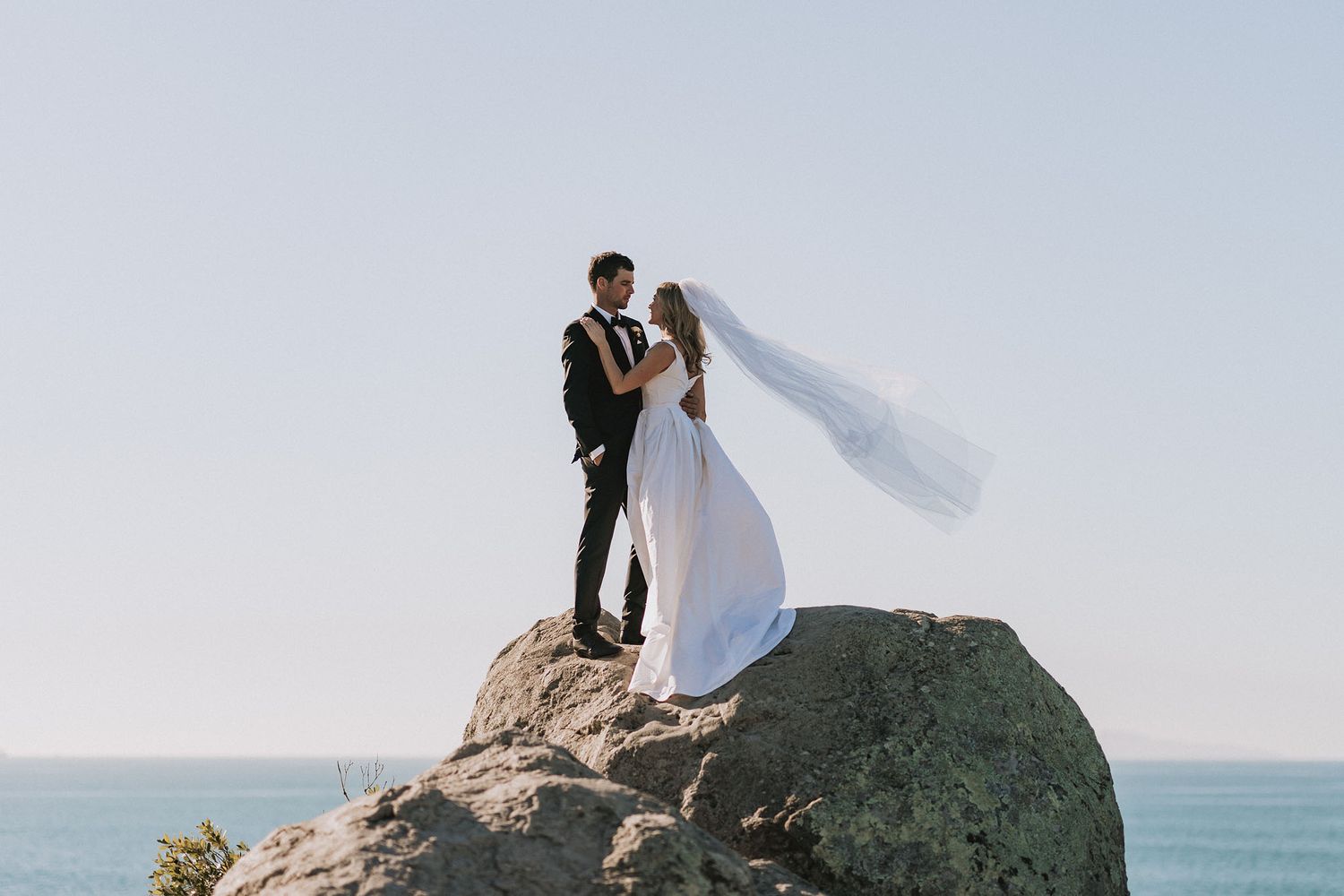 A couple in wedding attire stands together on a rocky cliff overlooking the ocean on their special day.