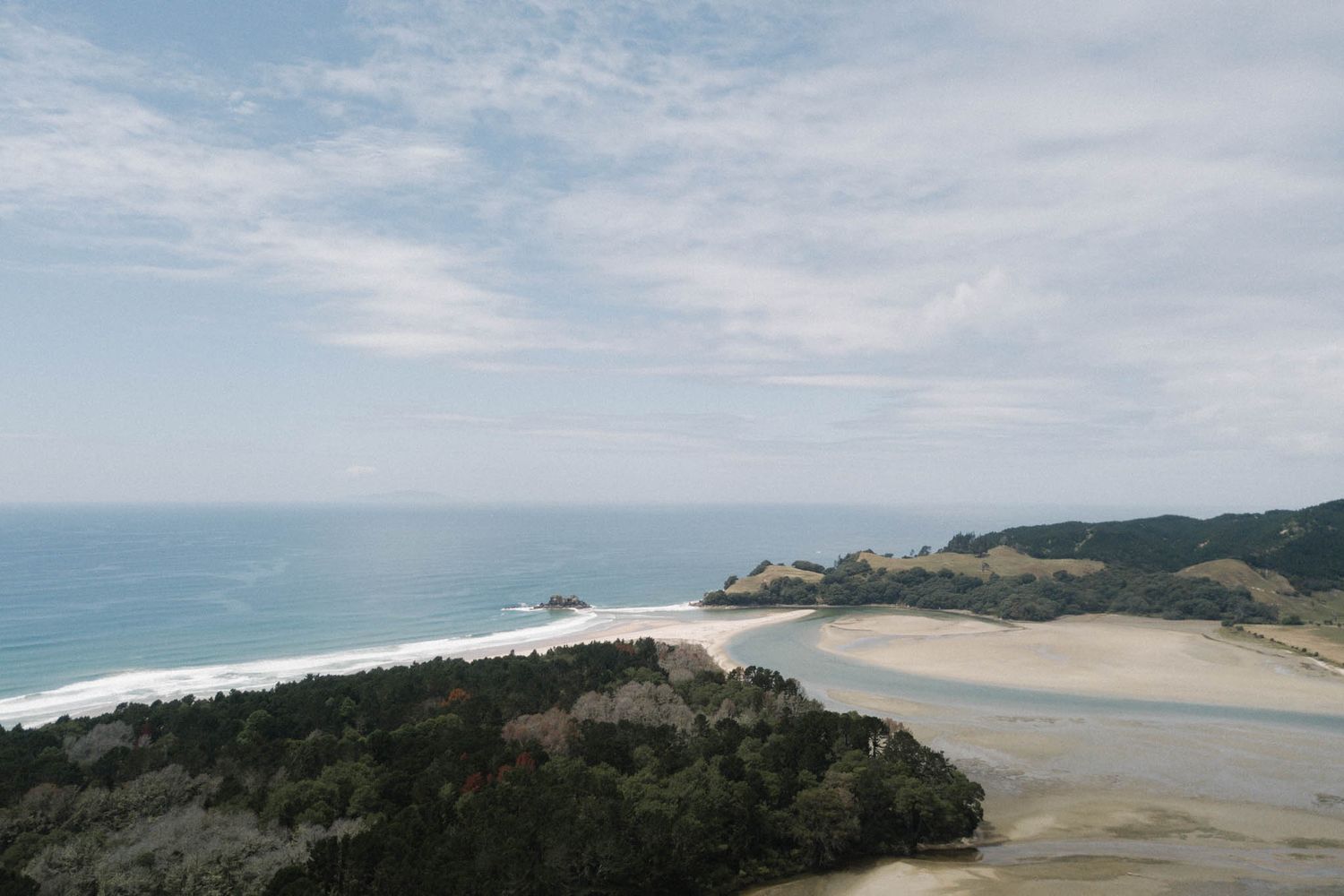 Scenic coastal landscape with sandy beach and turquoise waters meeting forested cliffs under light clouds.