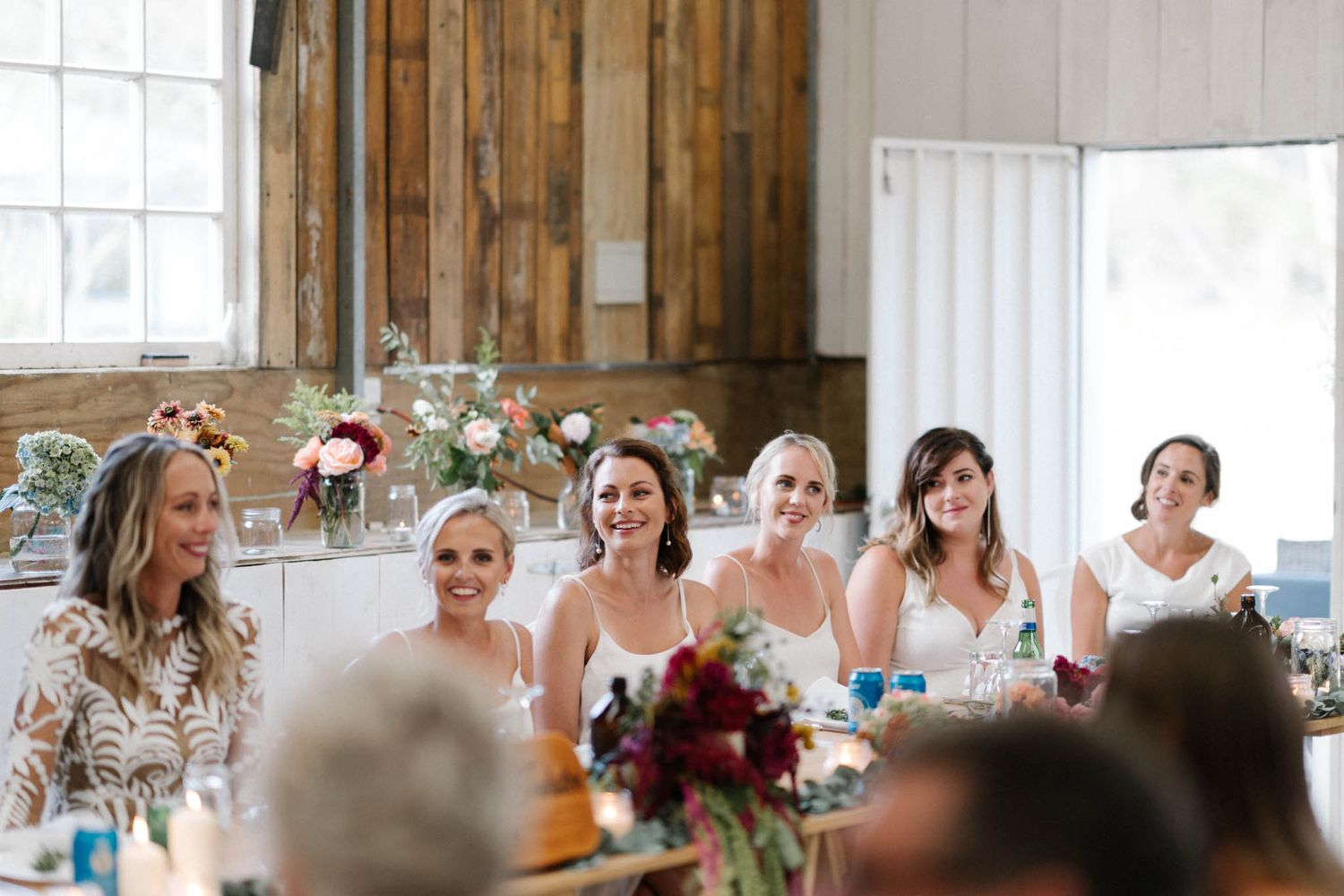 A group celebrates at an elegant reception table with rustic wood walls, floral centerpieces and natural light.