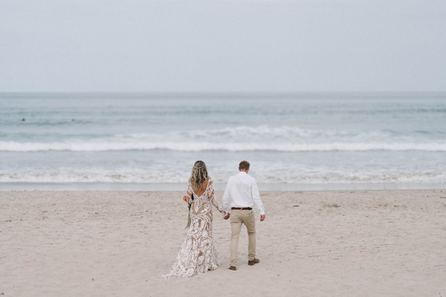 A couple in beachwear walks hand in hand along a misty sandy beach with ocean waves in the background.