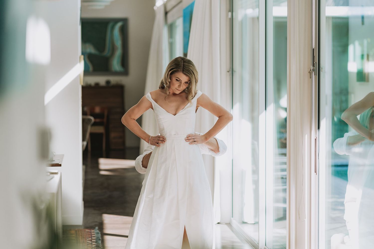 A white lace wedding dress hangs elegantly near a bright window in a light-filled room.