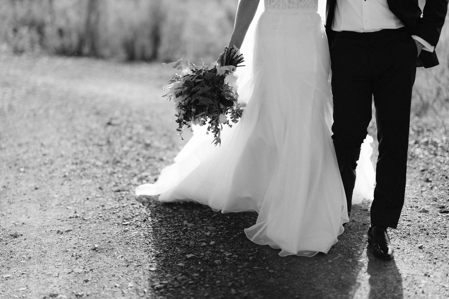 Black and white photo of a bride holding bouquet next to groom, showing just their formal wedding attire from waist down.