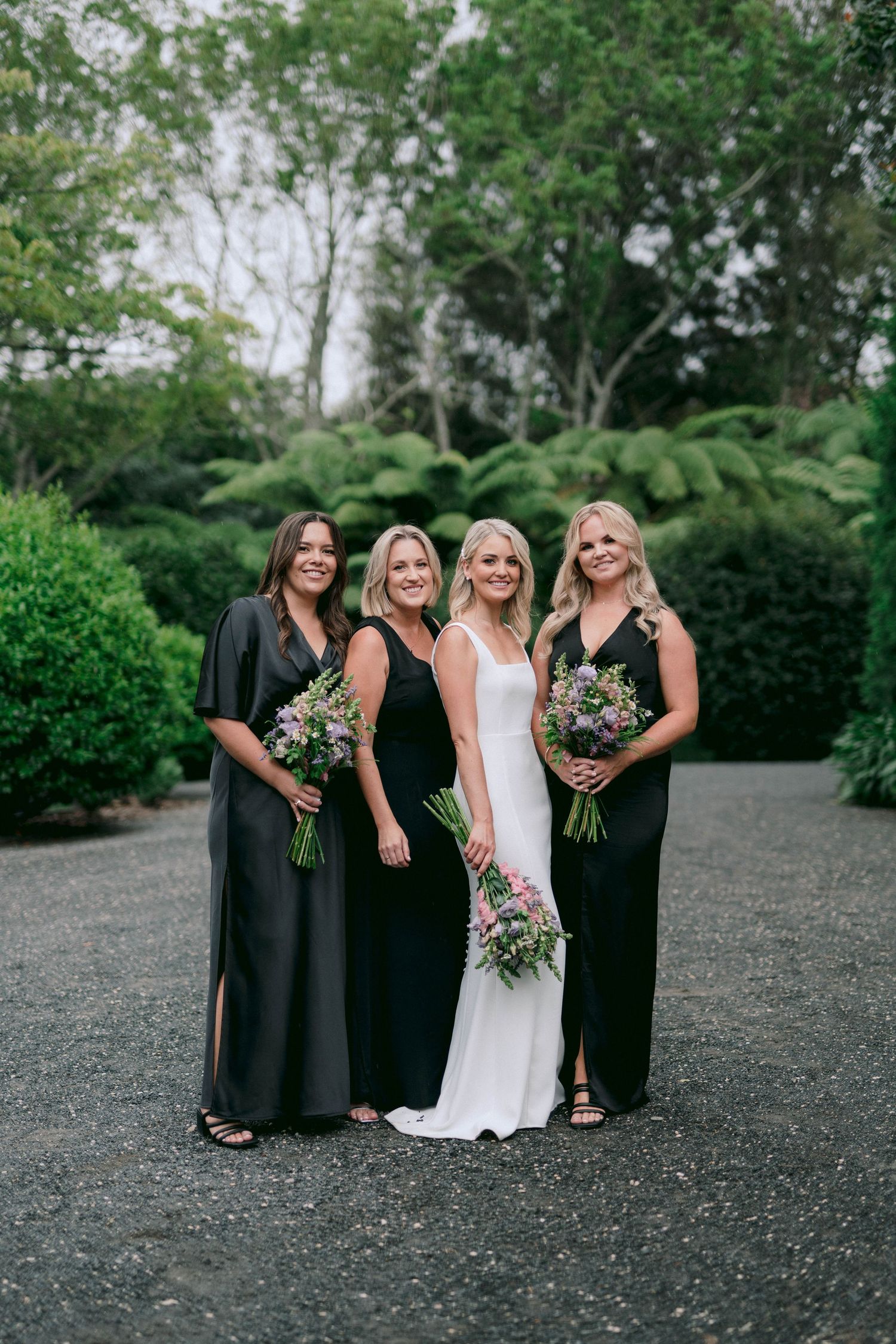 Wedding party posing outdoors with bridesmaids in dark dresses holding floral bouquets.