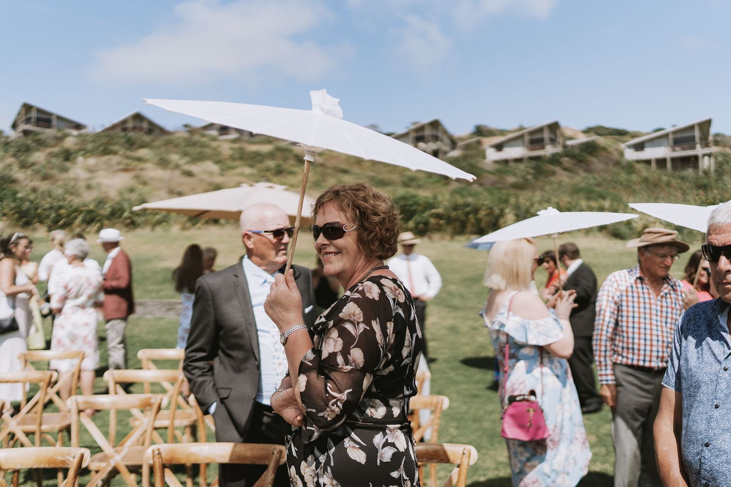Outdoor beach wedding guests mingling on grassy dunes with coastal houses in the background.