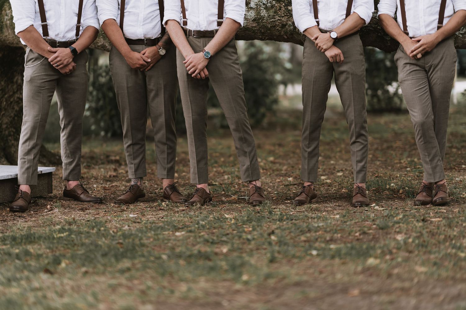 Row of groomsmen in khaki pants and white shirts stand outdoors with hands clasped in front for wedding photo.