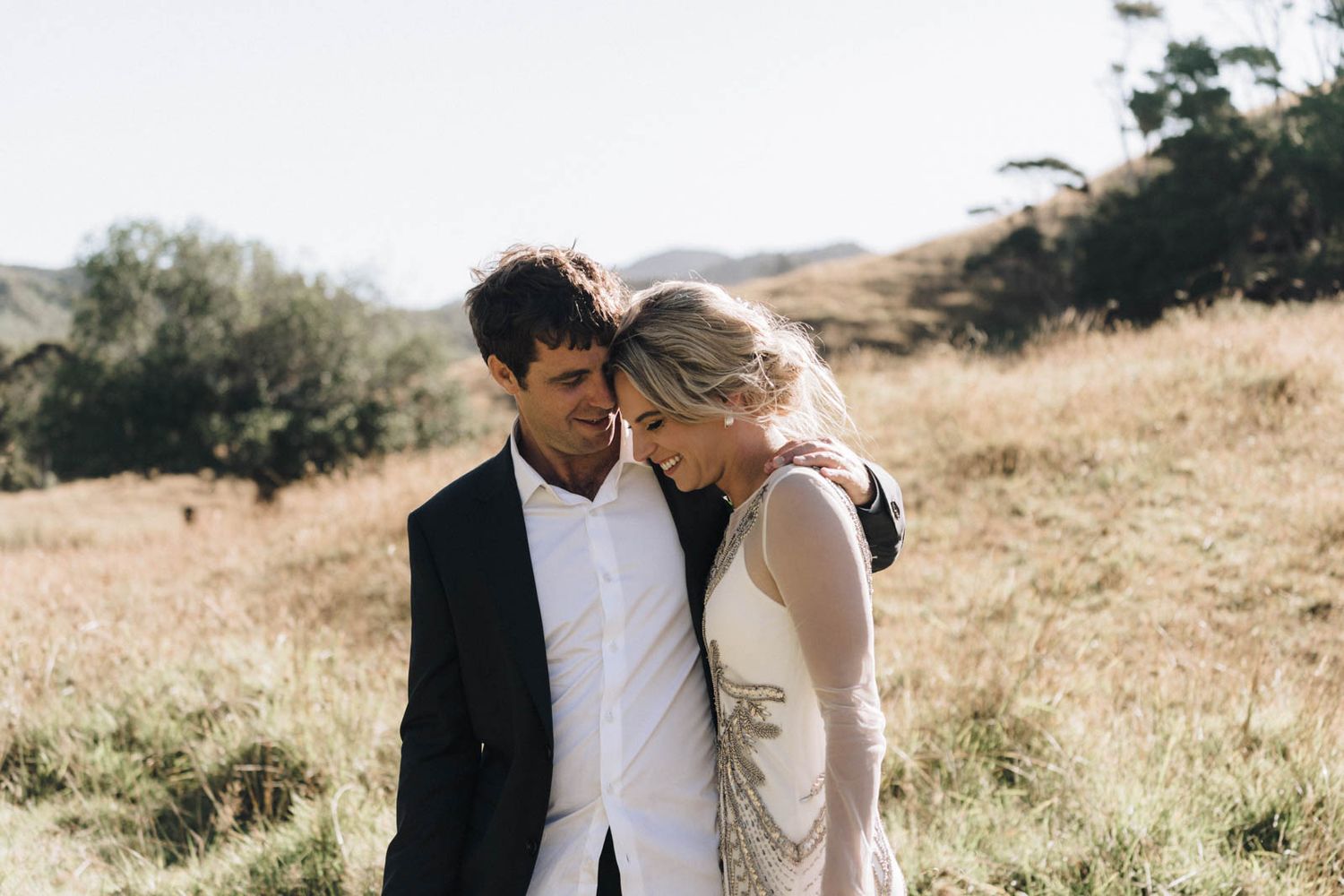 Couple in elegant wedding attire embraces lovingly in a sun-drenched golden meadow.