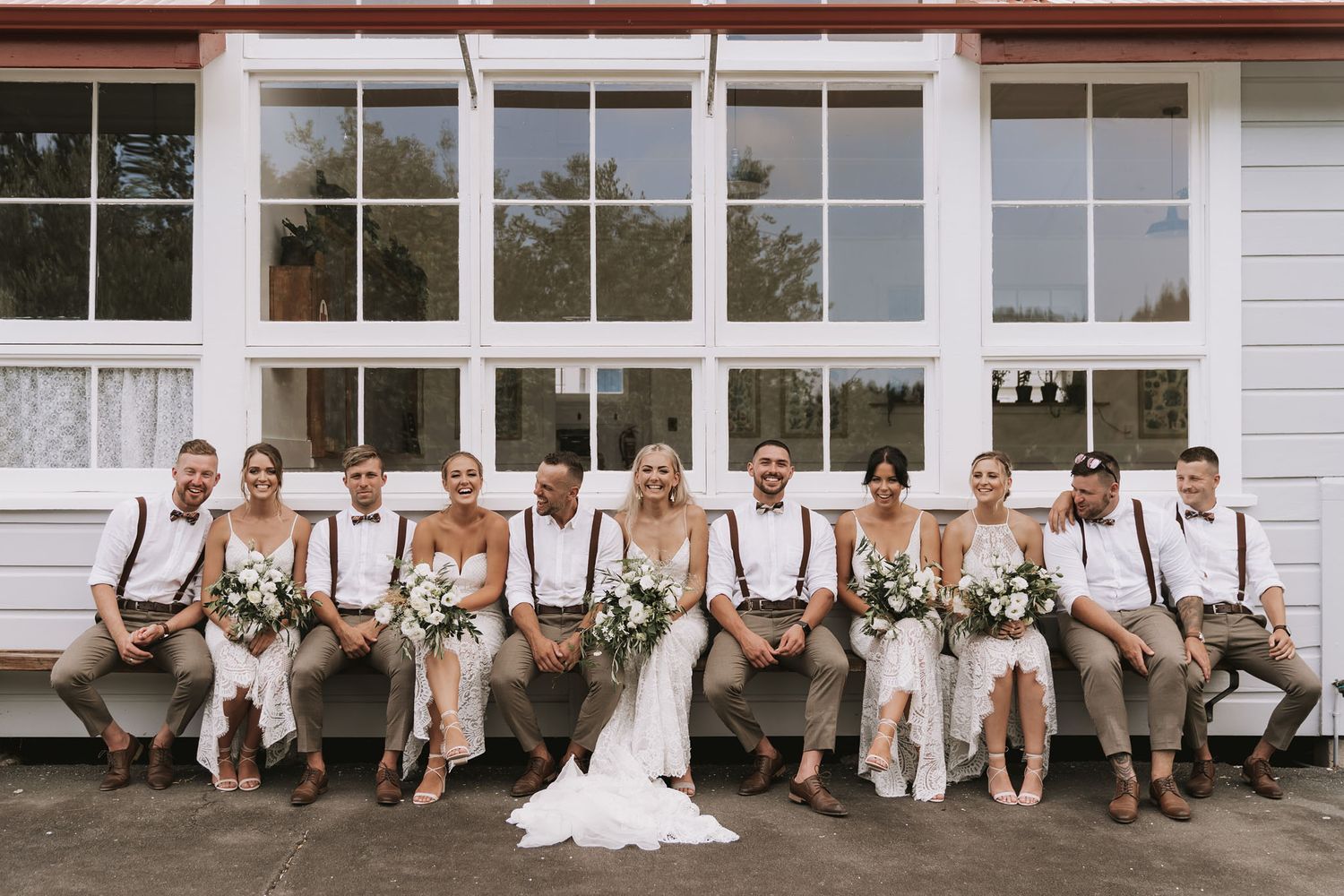 Wedding party in classic rustic attire with suspenders pose together on steps of white building porch.
