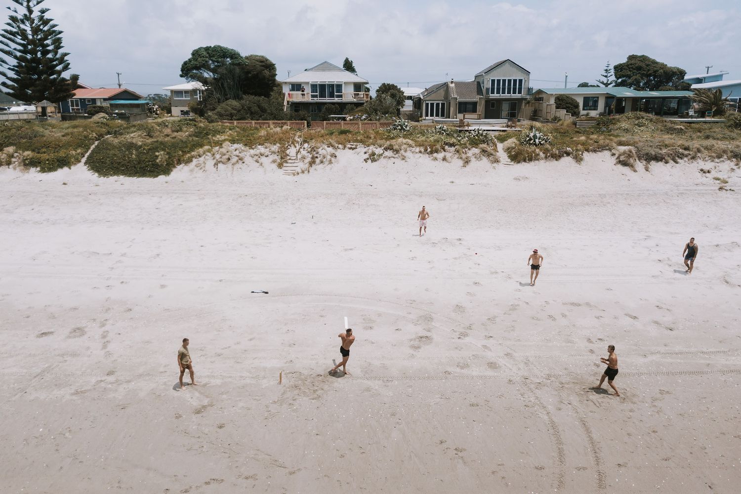 People walking on a white sandy beach with coastal homes and dunes visible in the background during a sunny day.