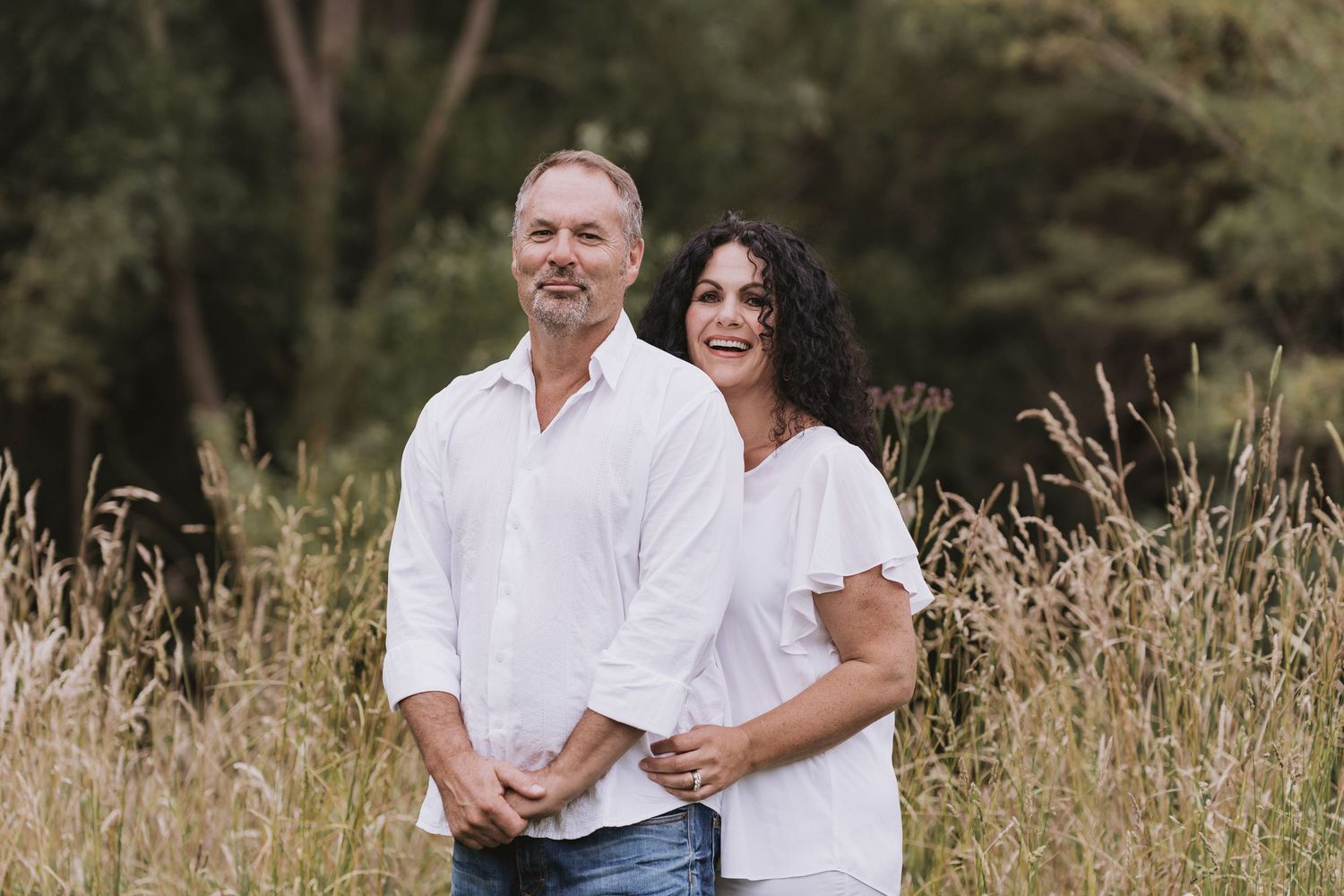 A couple in matching white shirts stands together in a field of tall autumn grass with trees in the background.