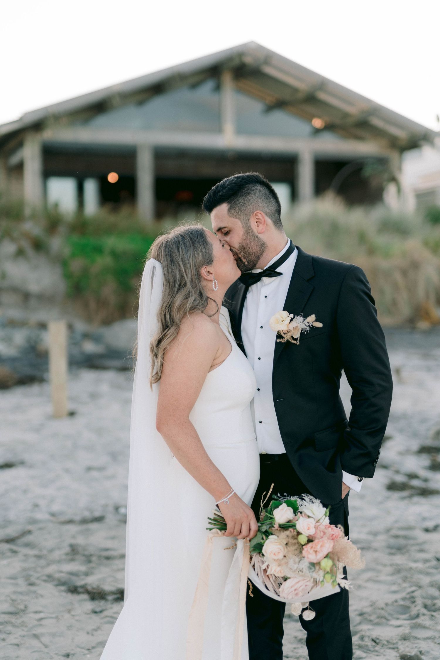 Couple enjoys romantic beach wedding moments at sunset with bouquet and elegant formal attire.