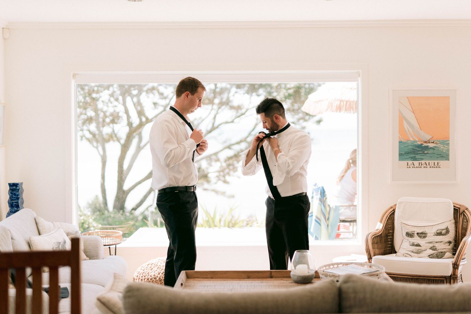 Two people in formal attire adjusting their ties by a large window in a bright living room.