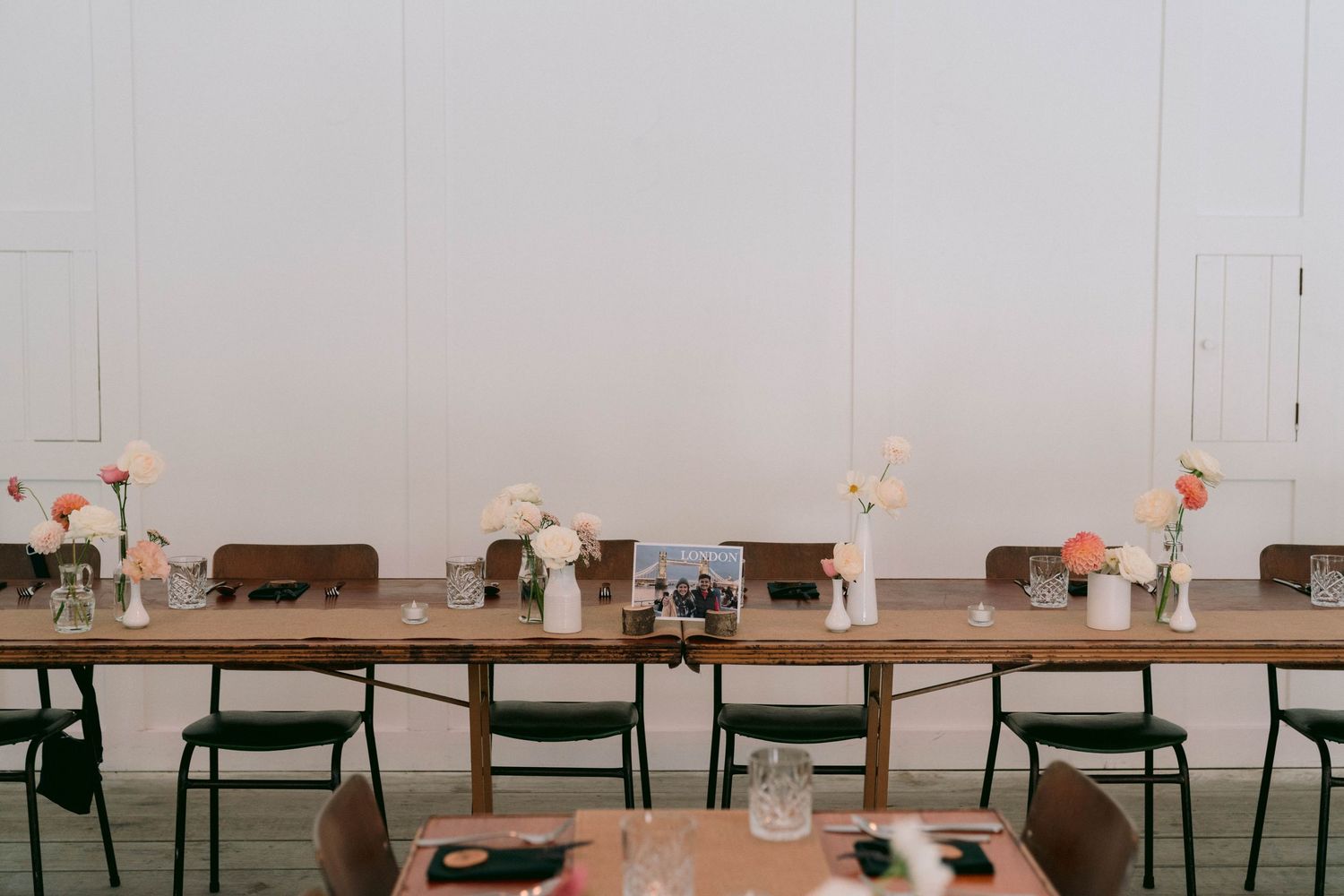 Long wooden dining tables set with simple floral centerpieces and black chairs in a minimalist reception space.
