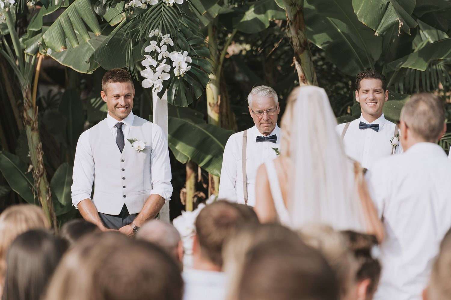 Outdoor wedding ceremony with attendees in white attire and tropical backdrop.
