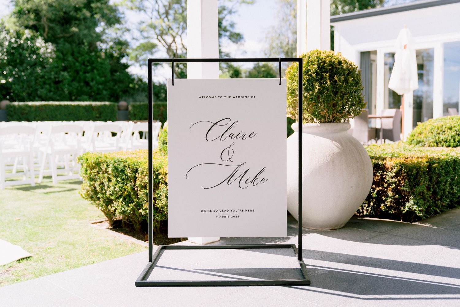 Elegant wedding welcome sign in black frame displayed on stand with greenery and white planters in background.