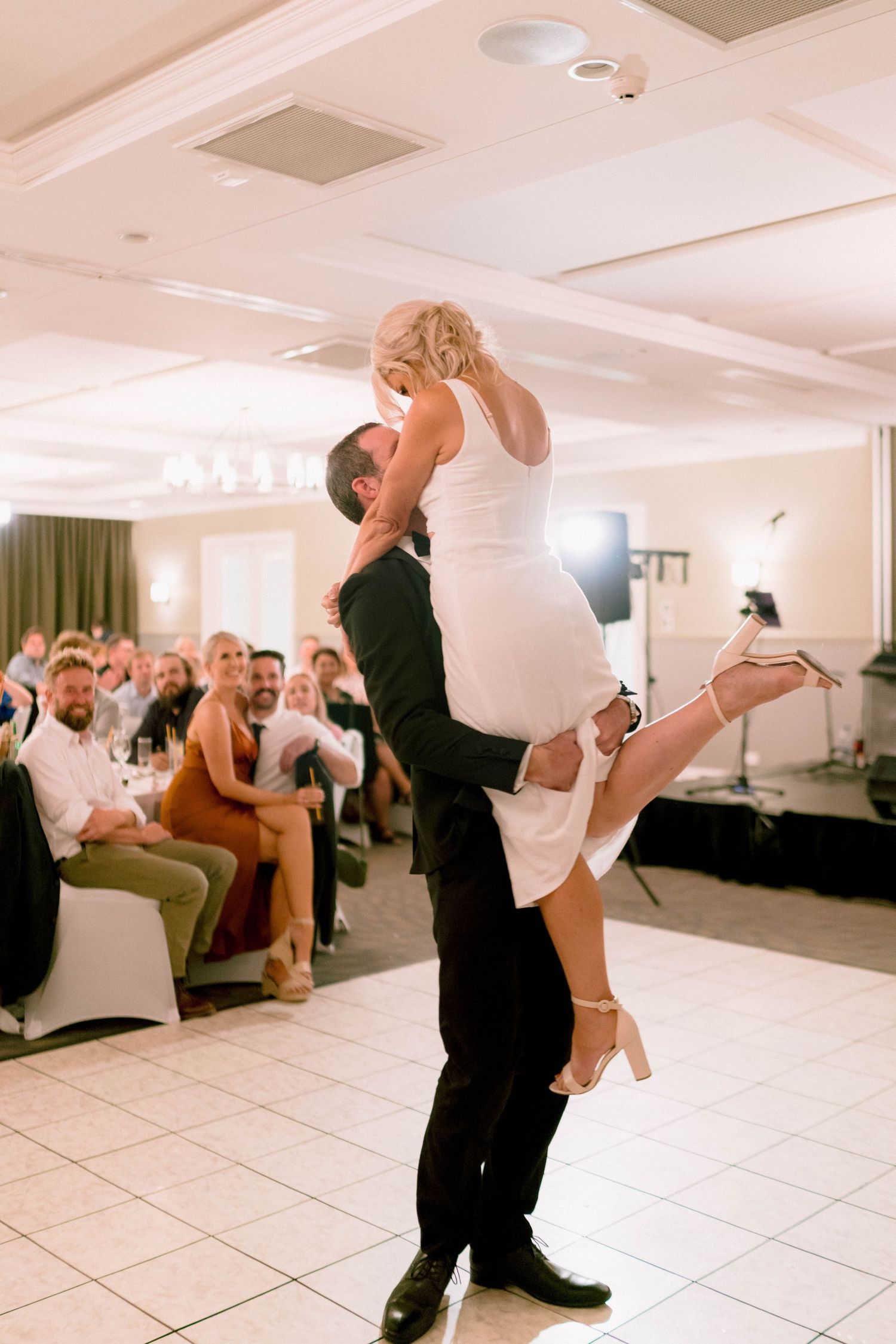 Wedding couple performs a dance lift move on a white tiled dance floor surrounded by guests watching.