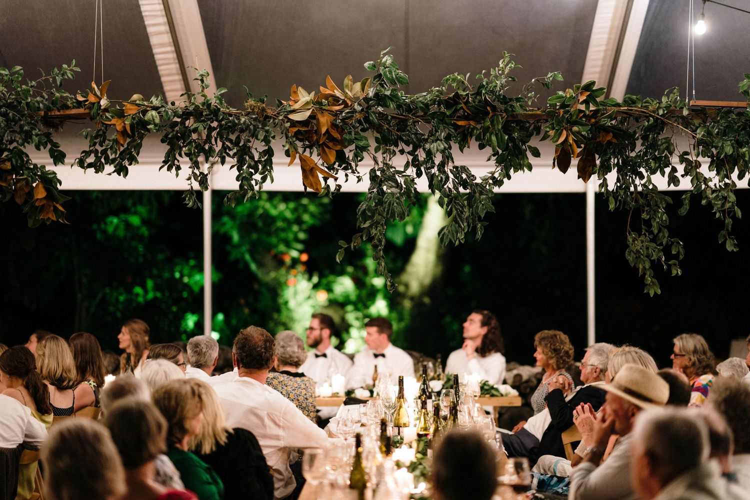 Evening wedding reception gathering with guests seated at illuminated tables beneath a transparent tent structure.