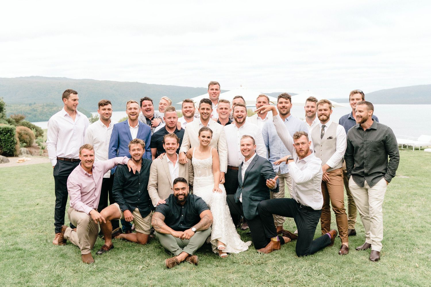 Wedding party gathered on a grassy hill overlooking a lake on a sunny day.