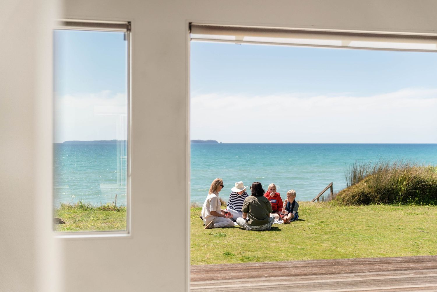 View through window frame of people sitting on coastal grass overlooking turquoise ocean and blue sky.