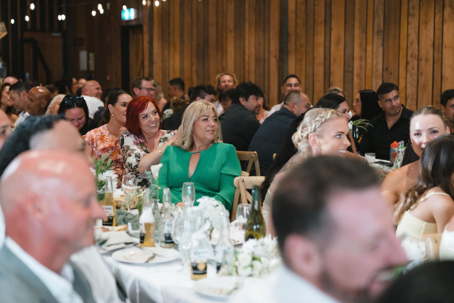 Wedding reception guests seated at tables in a banquet hall with wooden walls and ambient lighting.