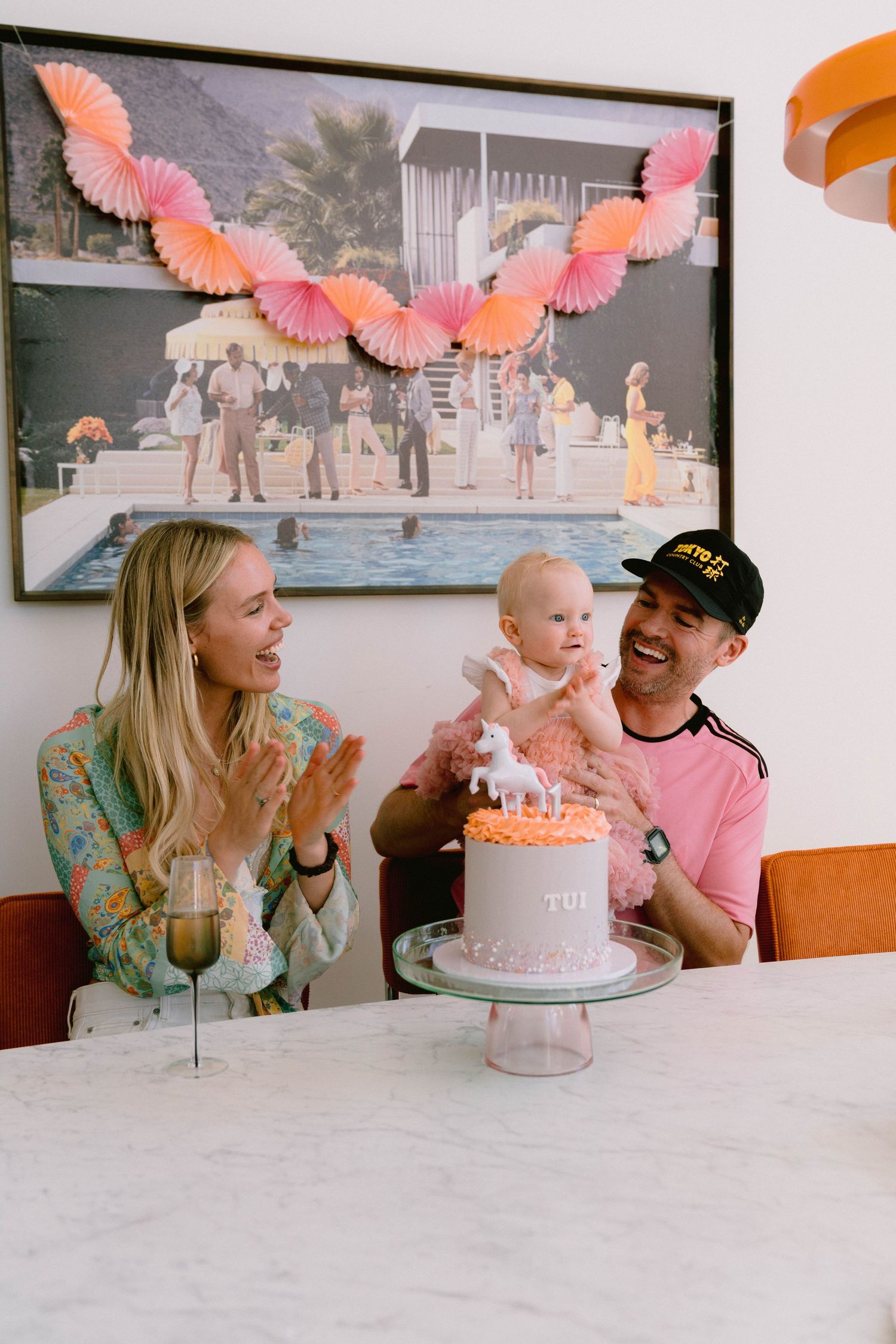 Family celebrating with a decorated birthday cake under pink paper fan decorations.
