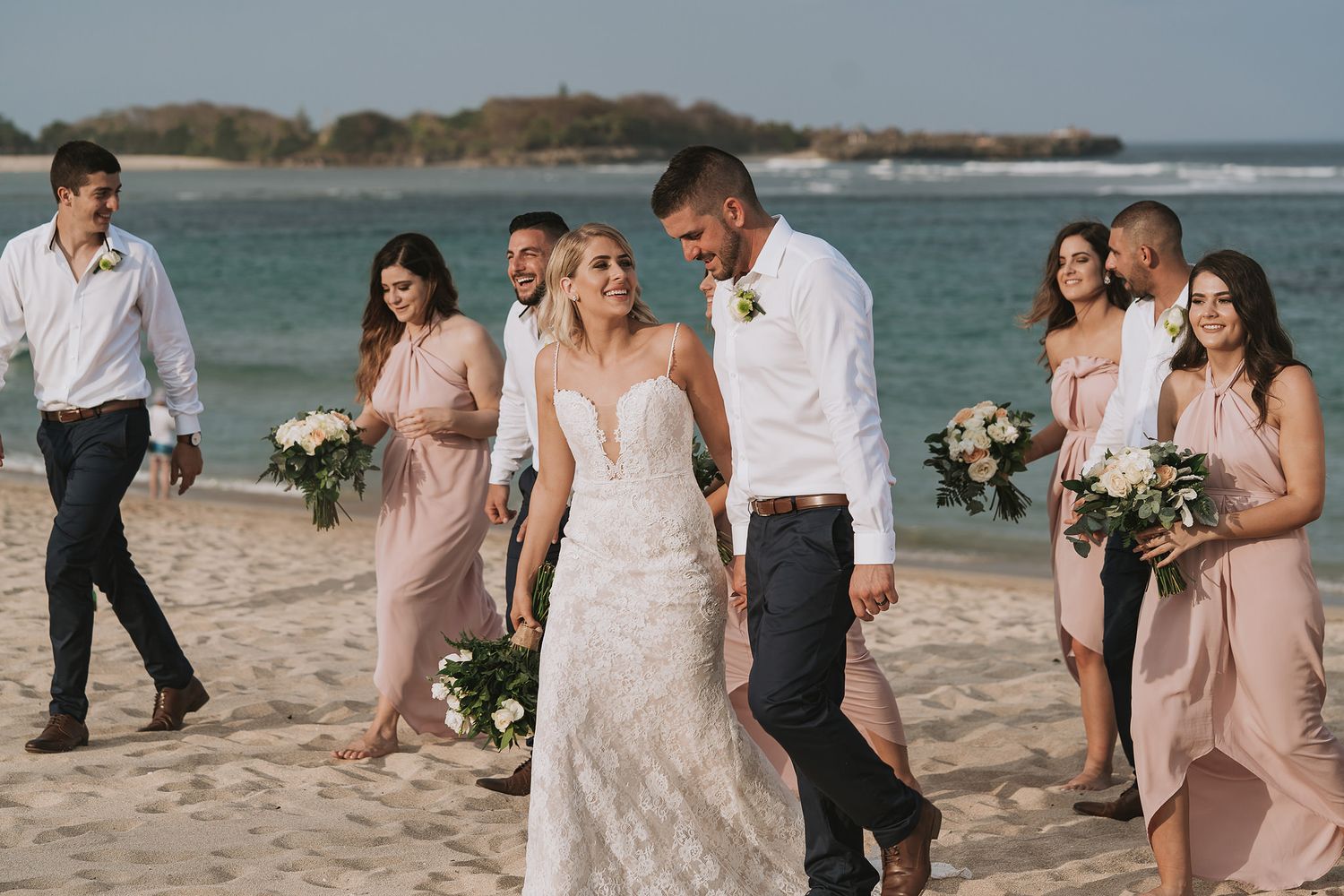 Wedding party walks along a tropical beach with turquoise waters in the background.
