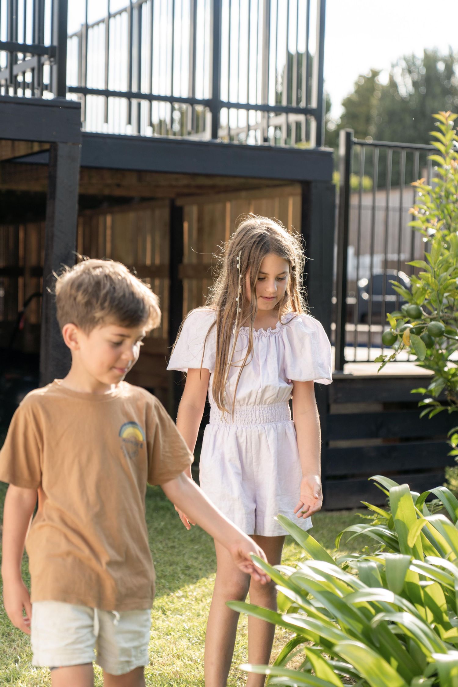 Children exploring lush garden plants along a black fence on a sunny day.