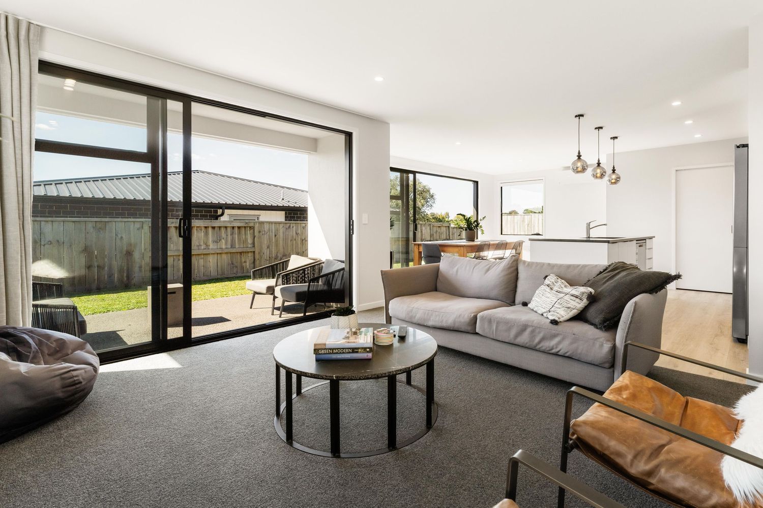 Modern living room with sliding glass doors, gray sofa, round coffee table and open concept design connecting to kitchen area.