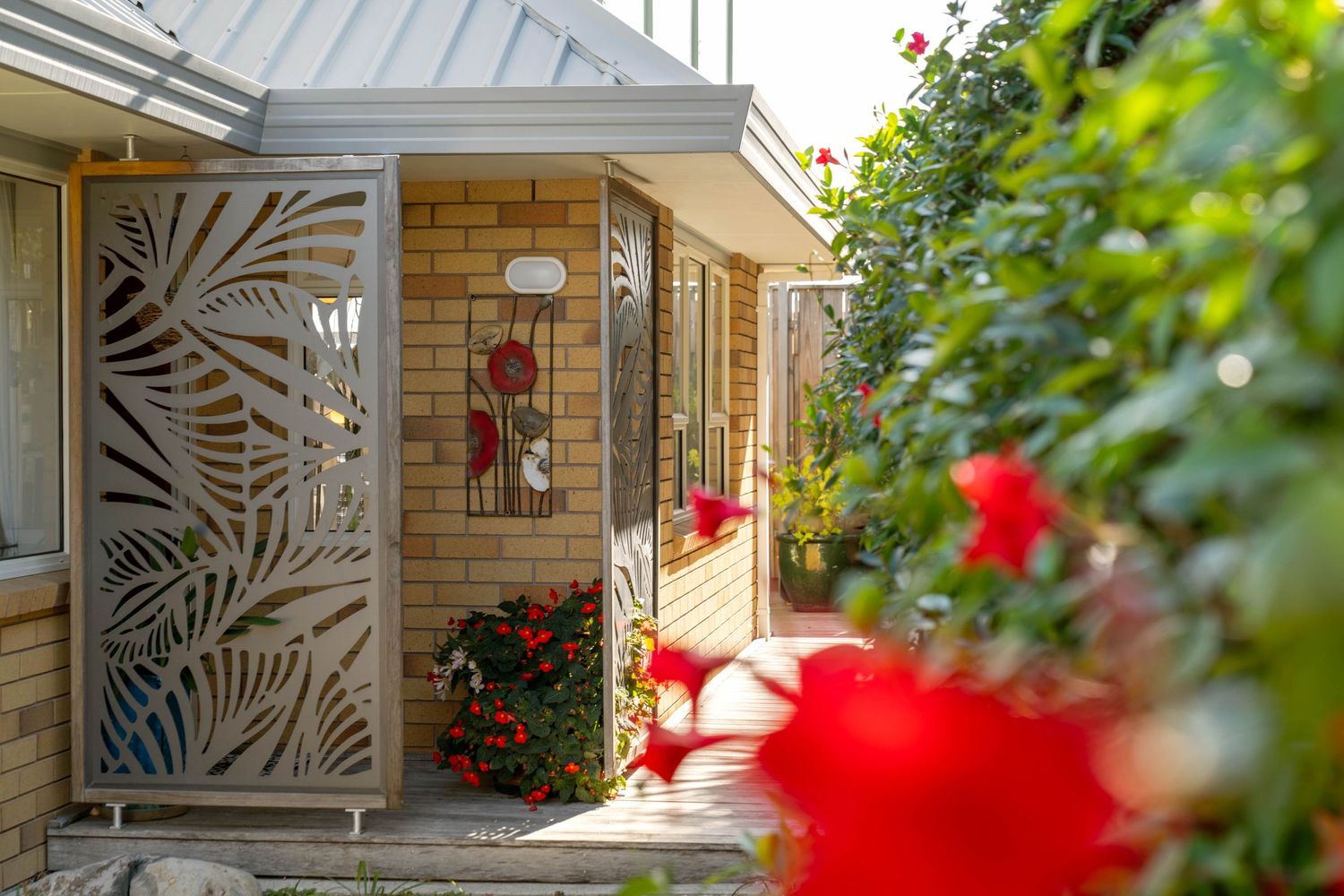 Decorative metal screen door with palm frond design leads to entryway with red flowers and greenery.