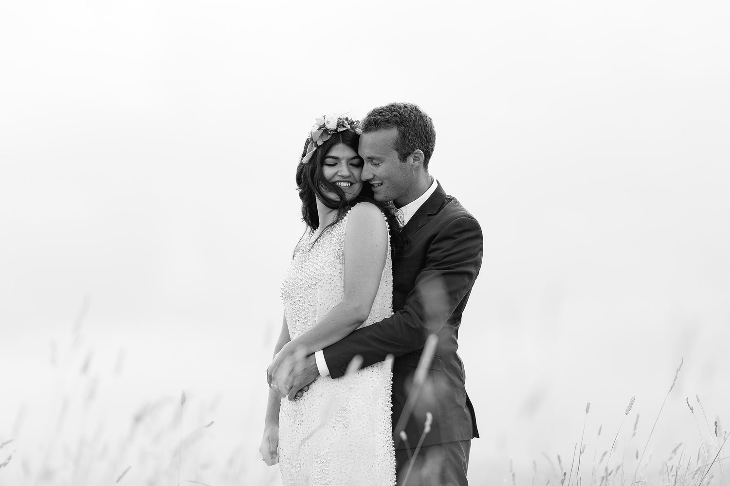 A romantic black and white wedding photo of a couple embracing in a field of tall grass against an overcast sky.