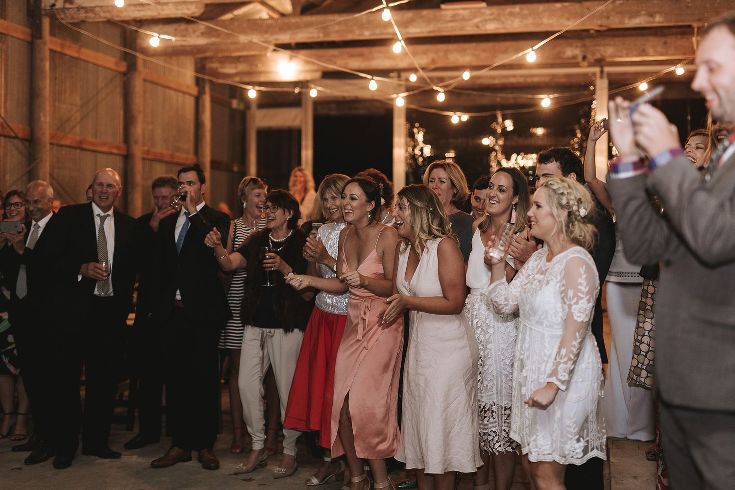 Wedding guests applaud and cheer during an evening reception in a rustic barn venue with warm lighting.