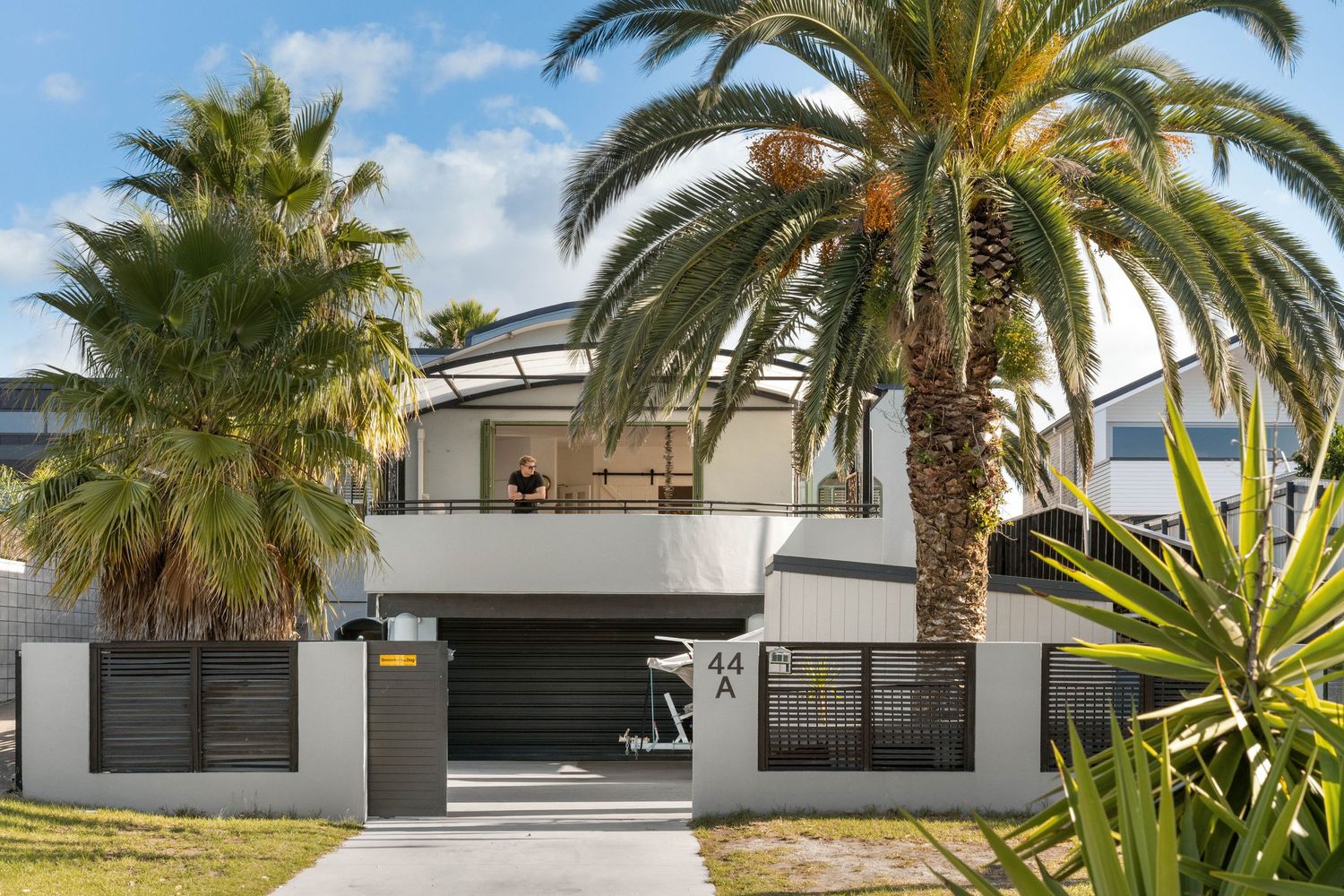Modern single-story home exterior with white walls, palm trees, and tropical landscaping framing the entry gates.