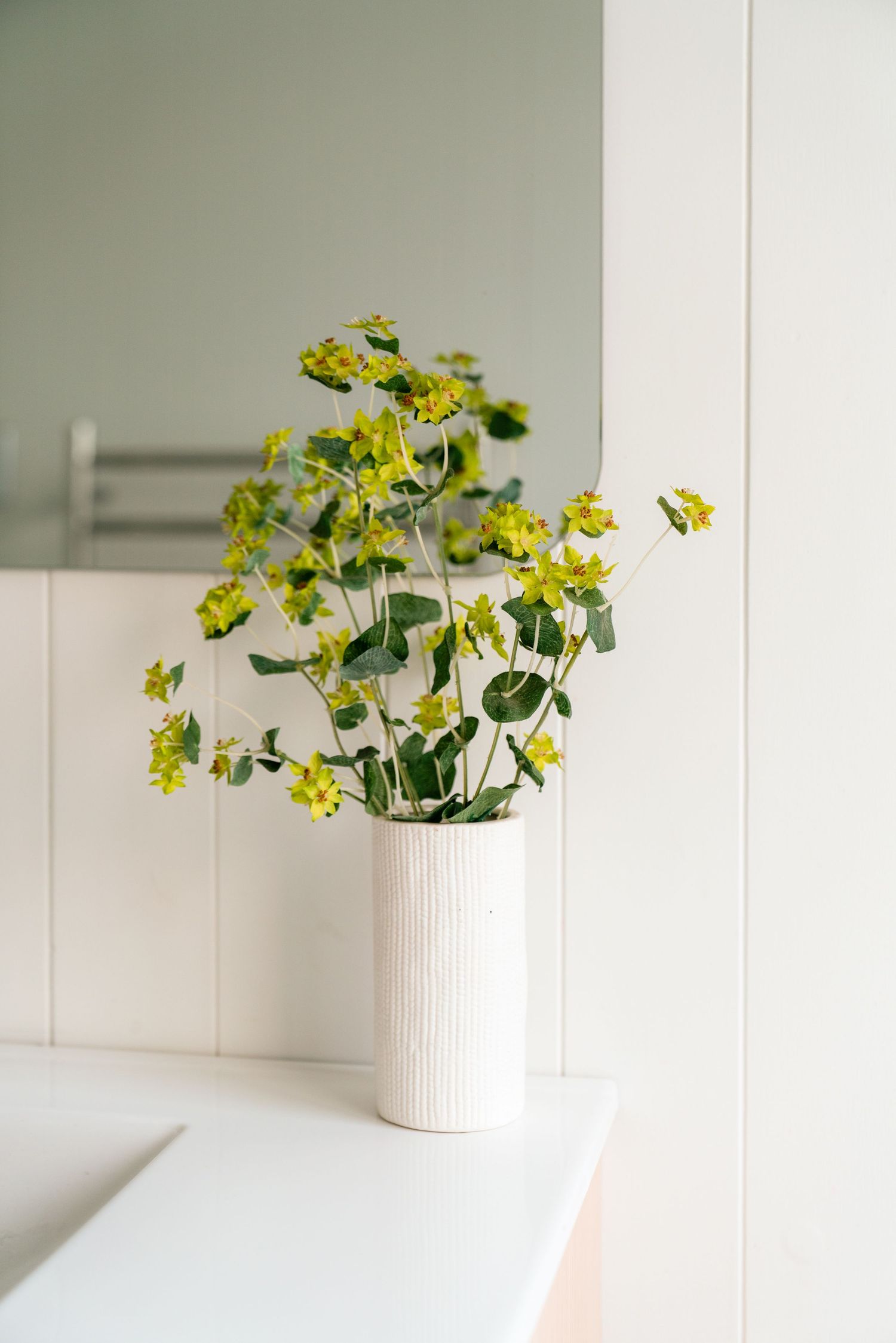 Yellow wildflowers arranged in a white ribbed ceramic vase against a light neutral wall.
